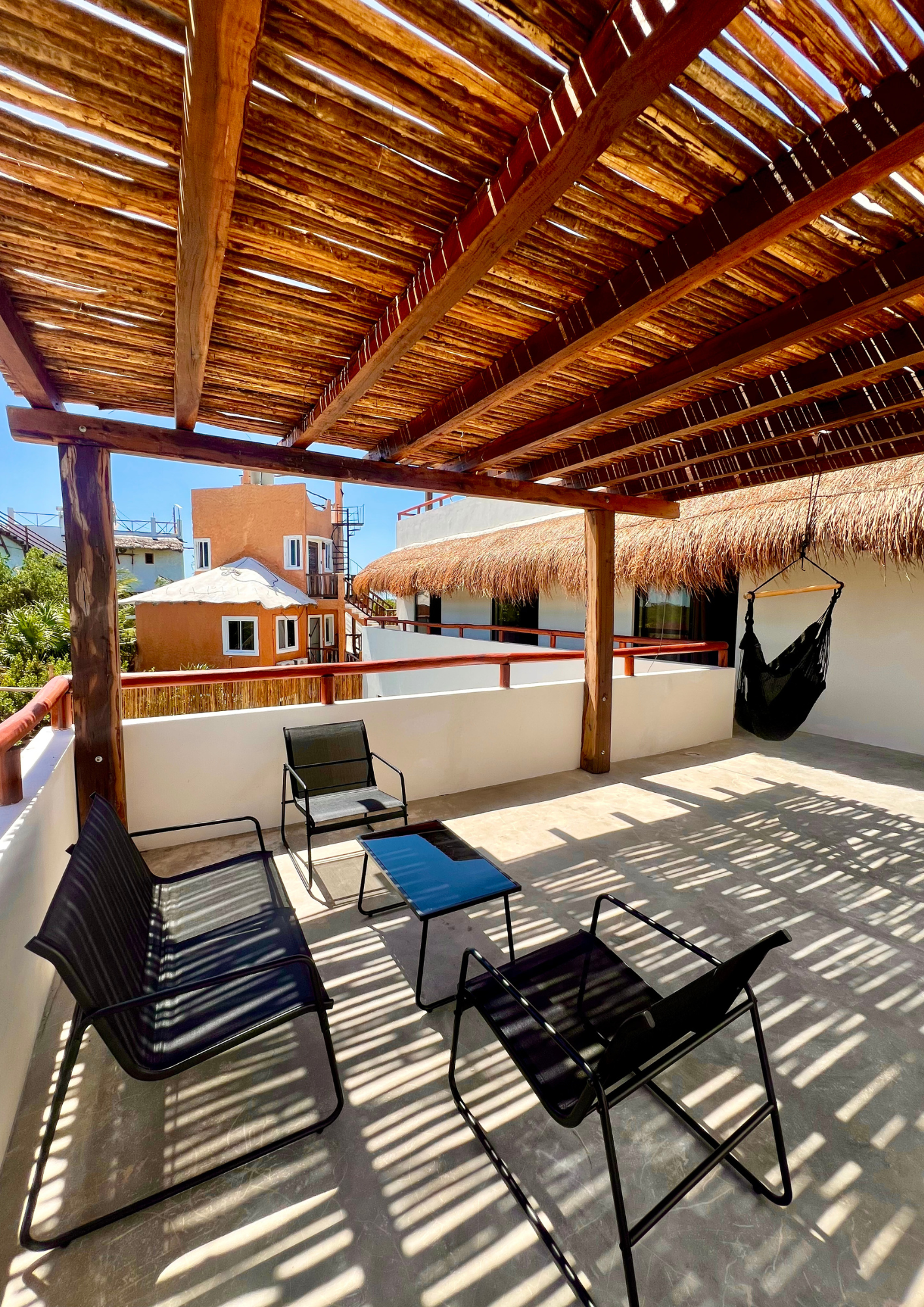 Terrasse ensoleillée avec mobilier noir, hamac, paravents en bambou, et vue sur des bâtiments colorés avec toits en chaume et ciel bleu.