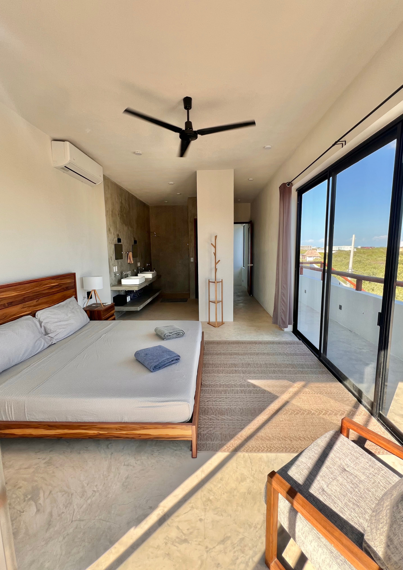 Chambre moderne avec un lit en bois, une grande fenêtre en verre, et une salle de bain attenante visible au fond.