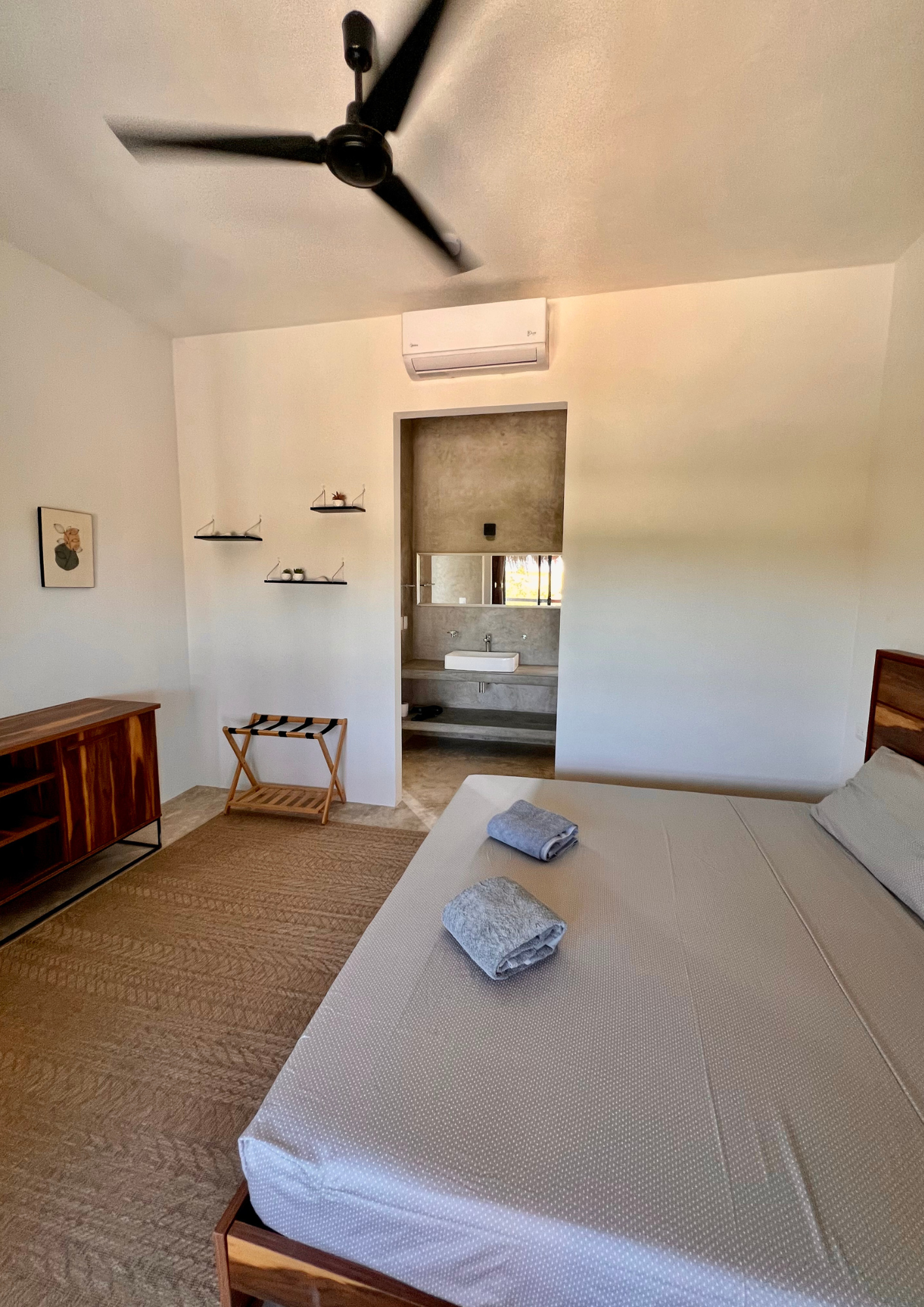 Chambre moderne avec lit, serviettes, bureau en bois, étagères et salle de bain attenante.