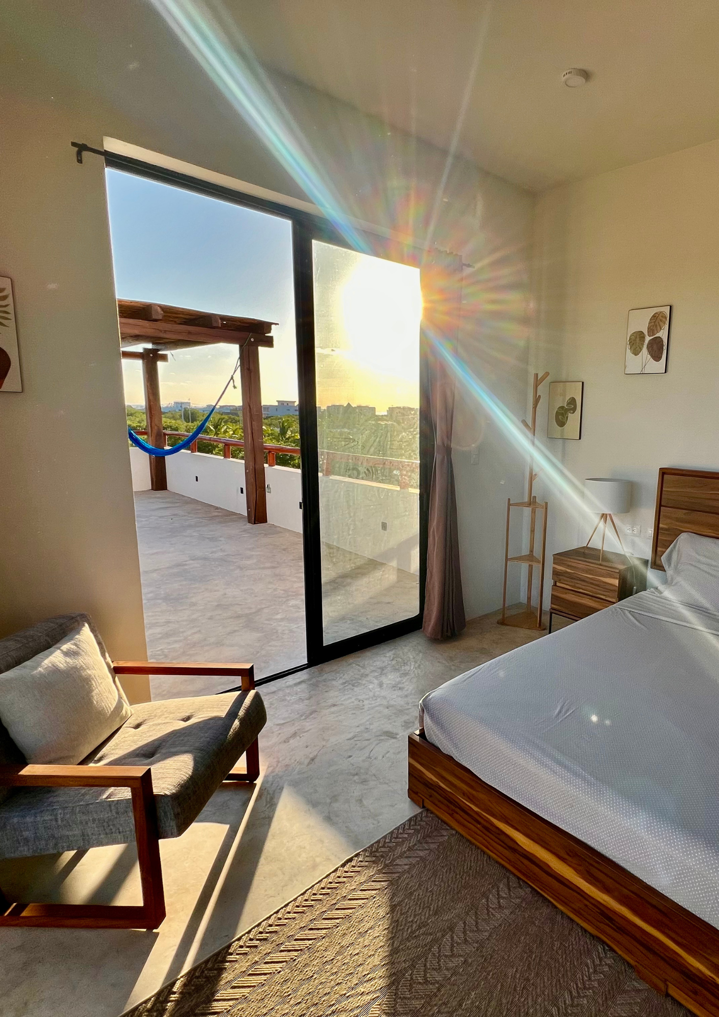 Chambre avec vue sur un balcon, soleil couchant, torii en bois avec hamac, mobilier en bois, tableau aux feuilles sur le mur, lumière naturelle