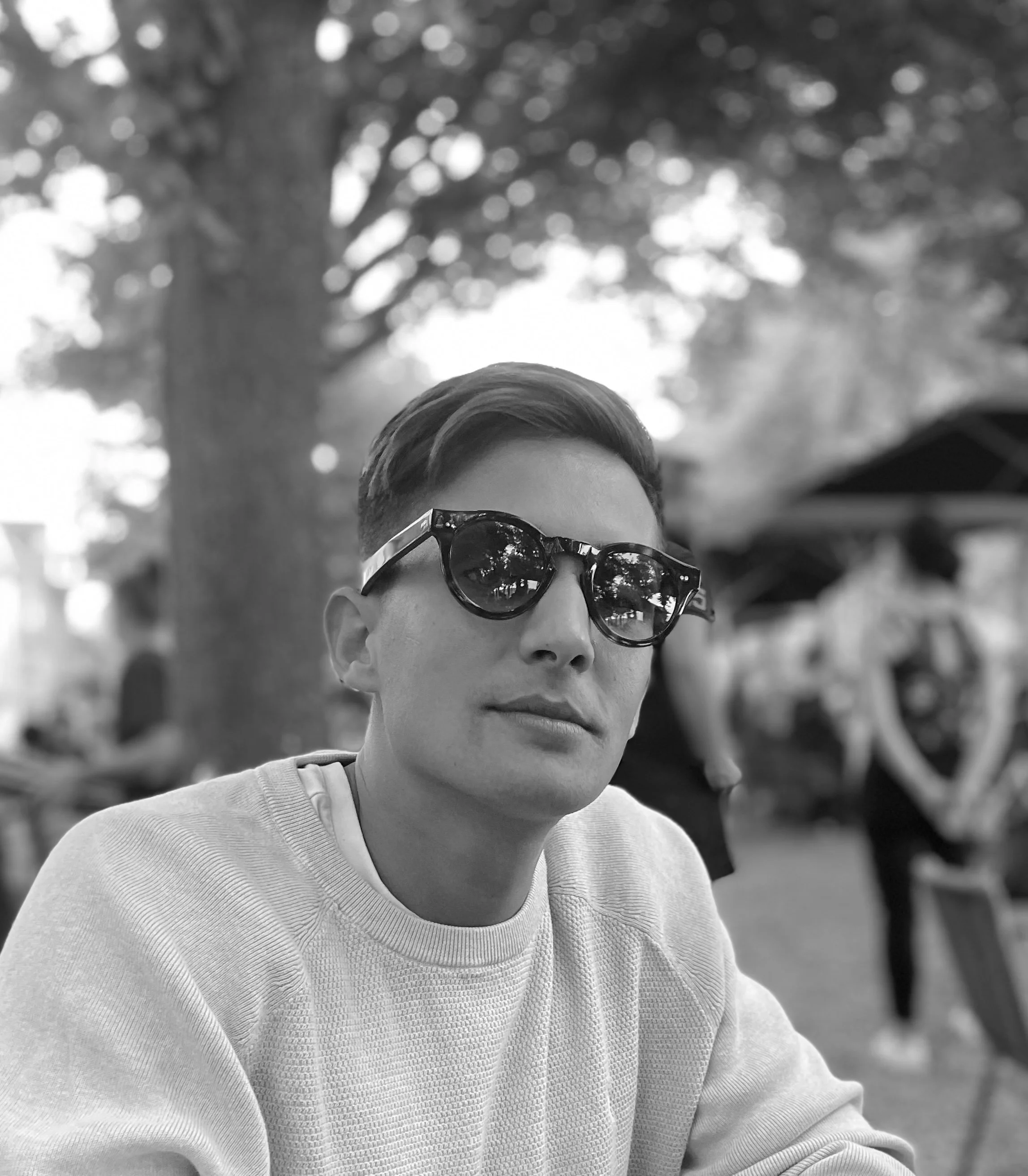 Un jeune homme portant des lunettes de soleil et un sweat blanc, assis à une table à l'extérieur avec des arbres en arrière-plan.