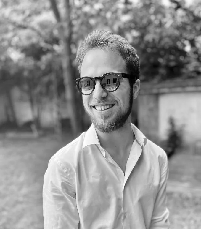 Un homme souriant porte des lunettes de soleil et une chemise blanche dans un jardin.