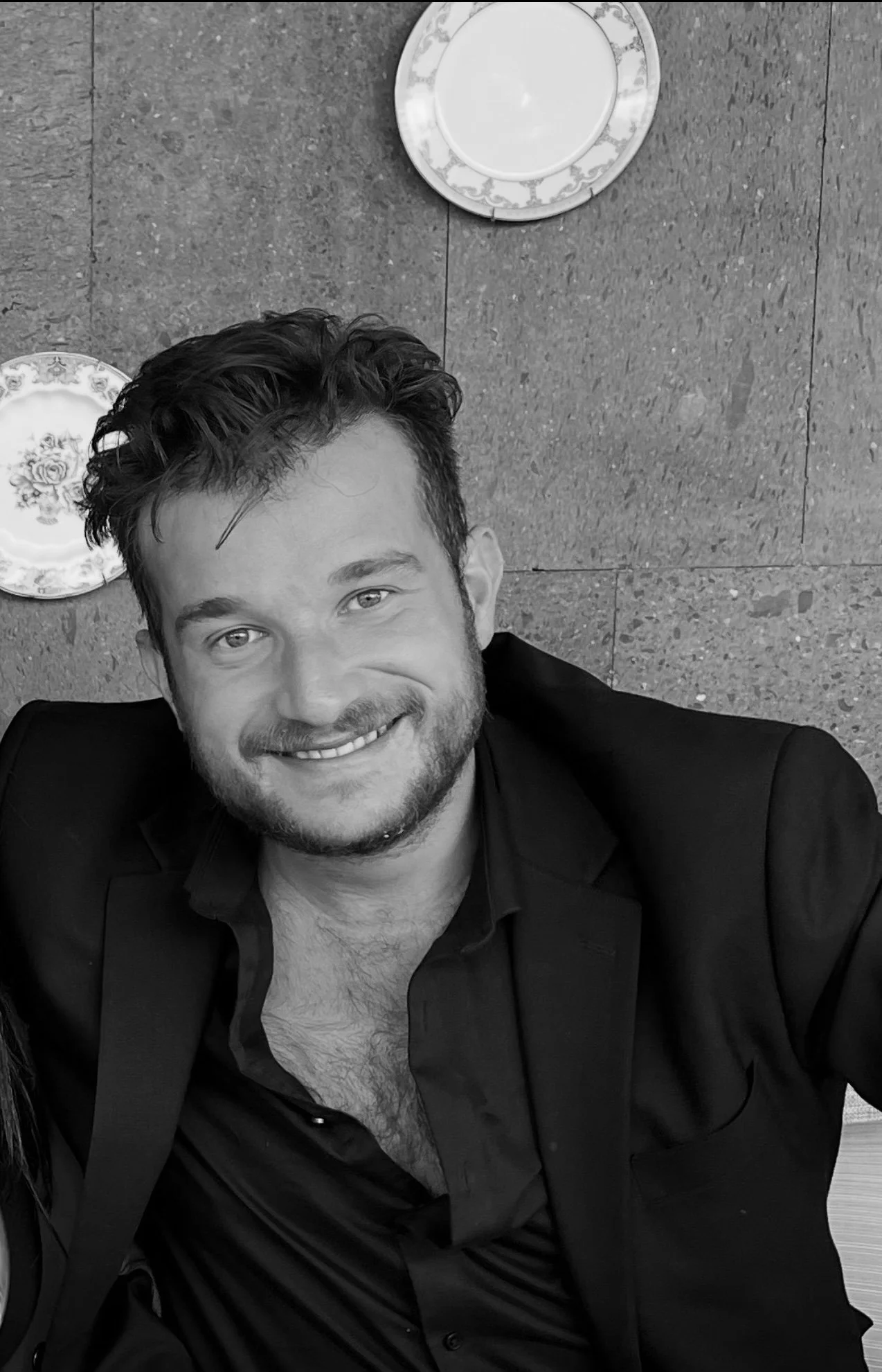 Un homme souriant, avec des cheveux bouclés et une barbe, vêtu d'une chemise noire et d'une veste, assis sur le sol en marbre, pris en photo en noir et blanc.