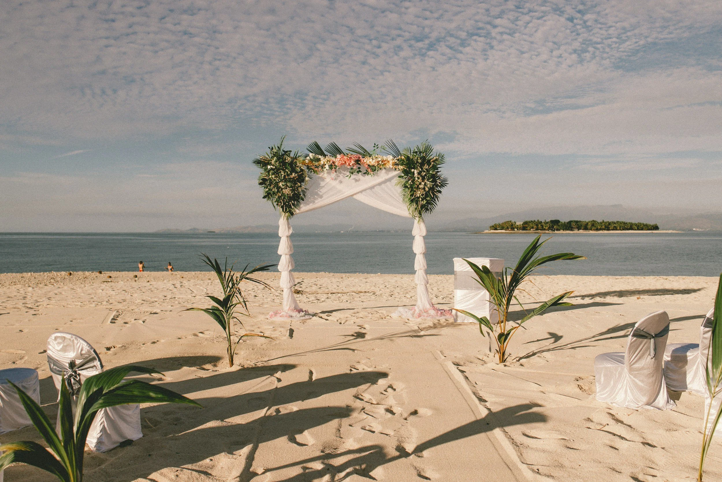 Alter de mariage décoré avec des fleurs, situé sur une plage en bord de mer, avec des chaises couvertes de tissu blanc et des plantes vertes