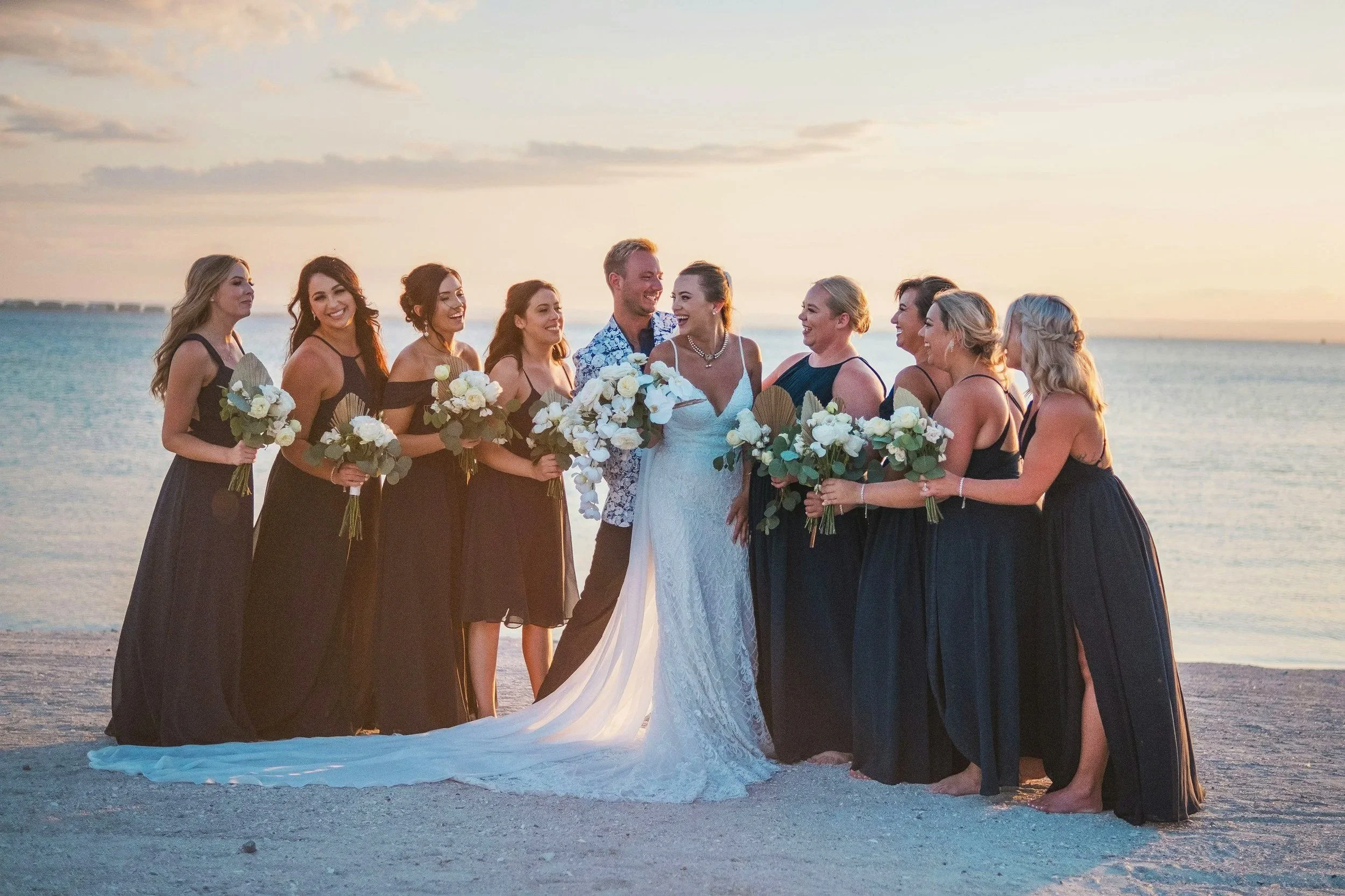 Un groupe de personnes, dont une mariée en robe blanche, se tient sur la plage au coucher du soleil, entouré de femmes en robes longues et de bouquets de fleurs.