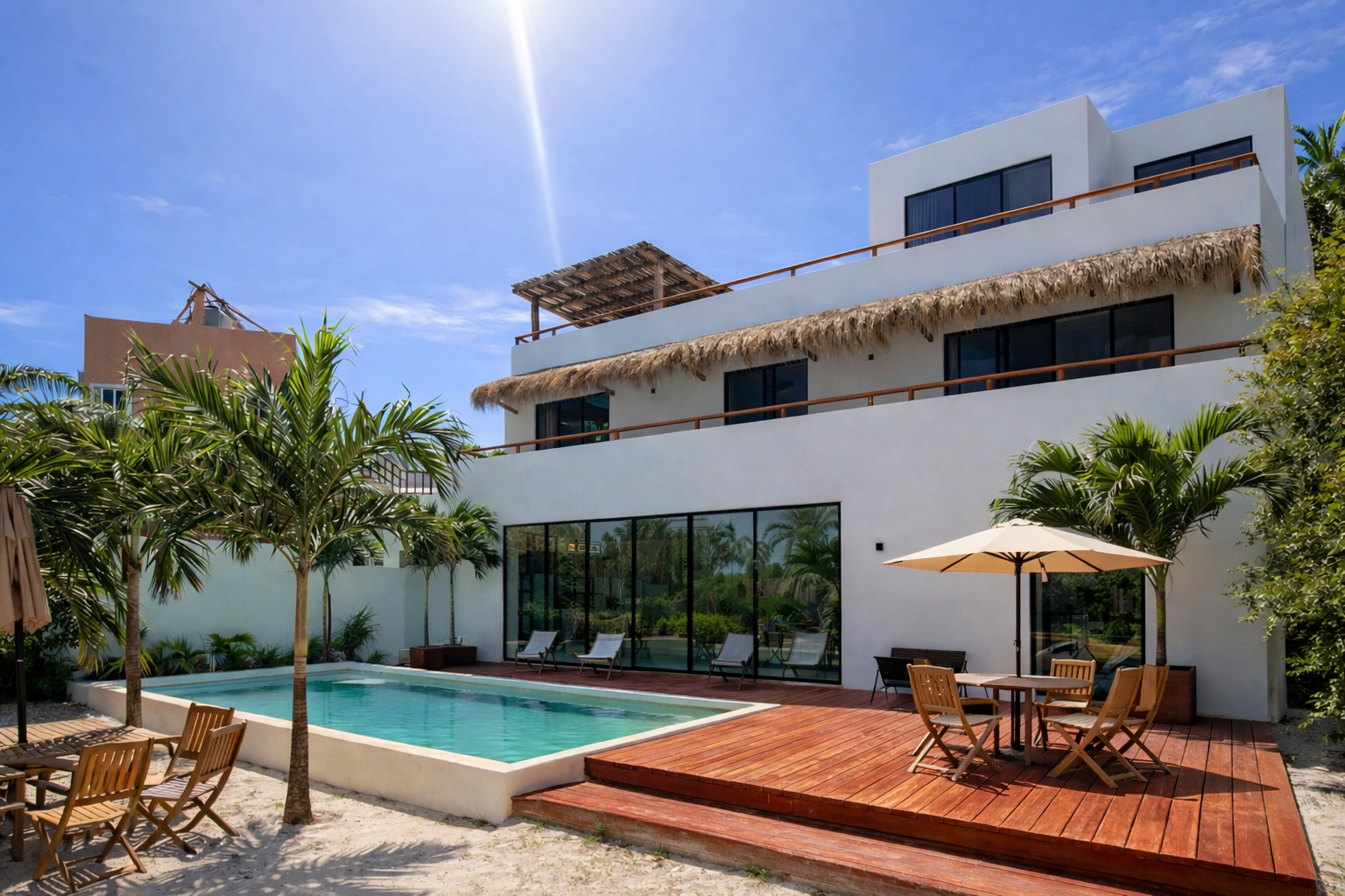 Une maison moderne à plusieurs étages avec une piscine et une terrasse en bois, entourée de palmiers, sous un ciel ensoleillé.