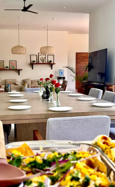 Salle à manger moderne avec une grande table en bois, ornée de plusieurs vases avec des roses rouges, et une télévision à écran plat sur le mur à droite. En arrière-plan, un salon avec un canapé beige, des étagères décoratives, et un environnement lu