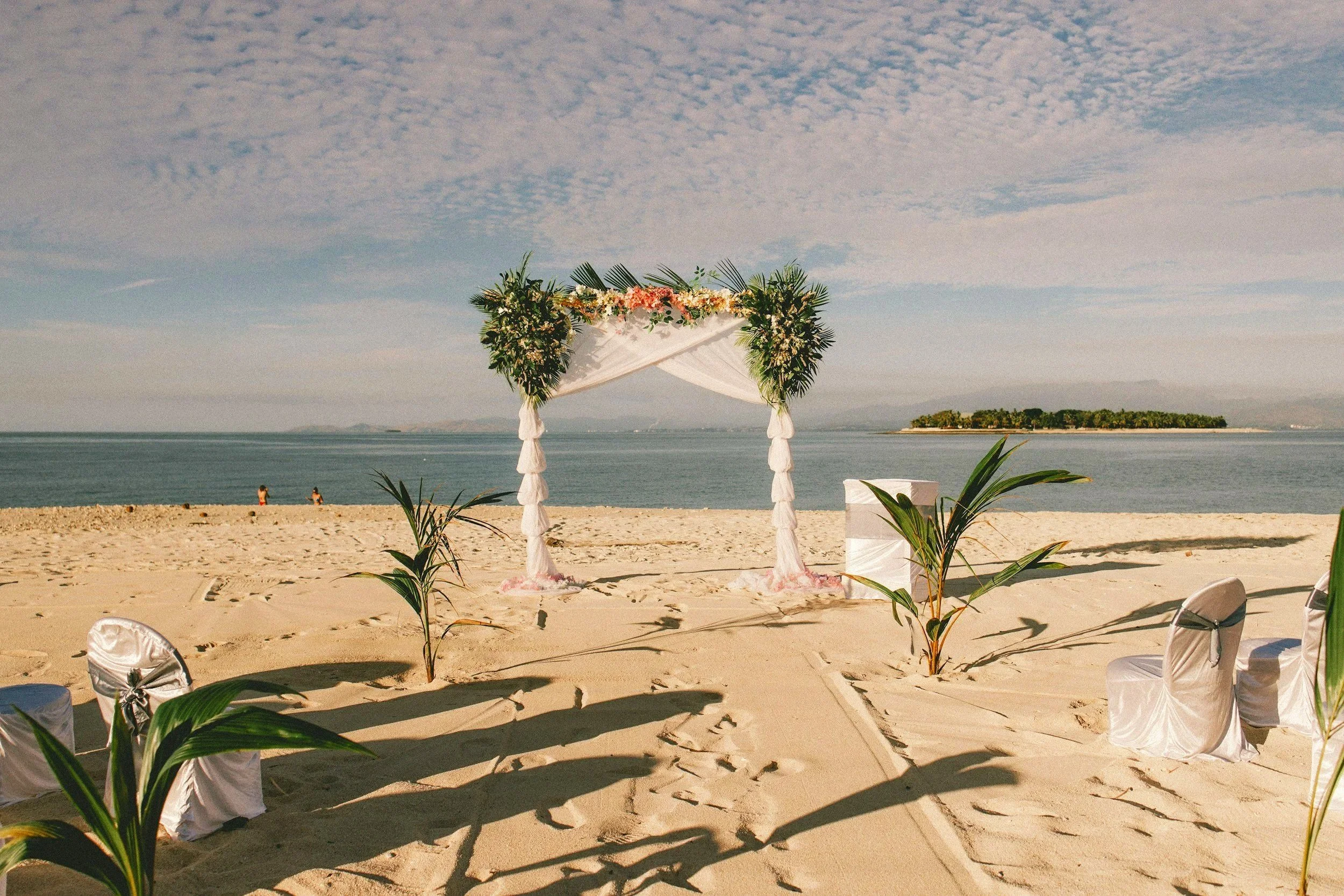 Décor de mariage sur la plage avec un arc floral, chaises blanches couvertes de satin, et des plantes tropicales, avec la mer et une petite île en arrière-plan.
