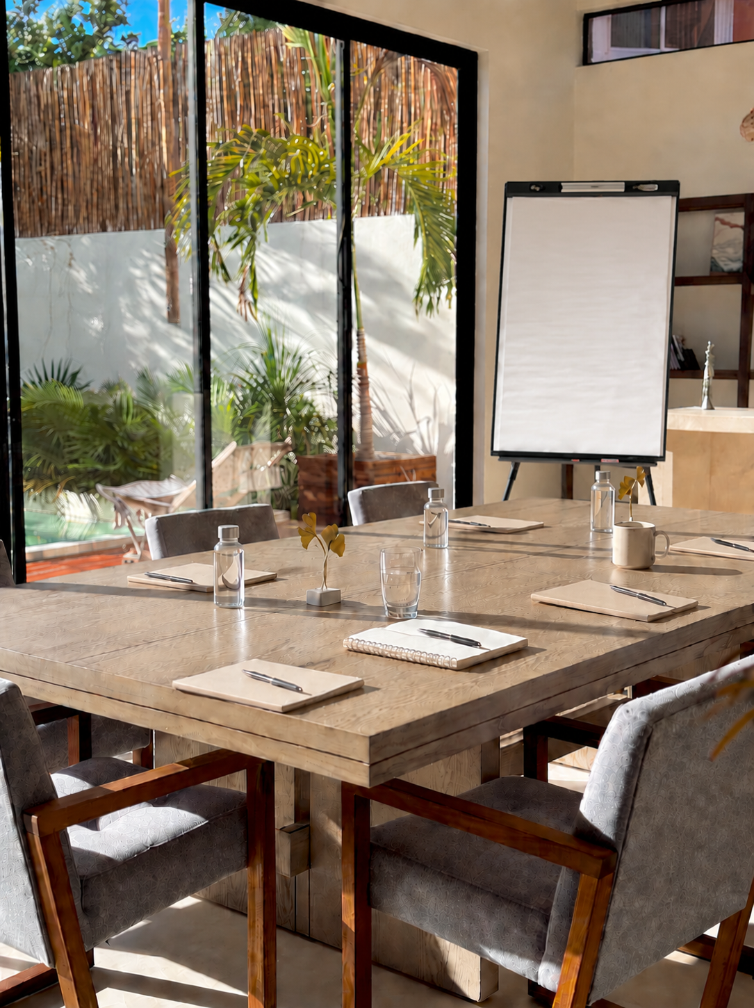 Salle de réunion avec une grande table en bois, des chaises, des bouteilles d'eau, des tasses, des blocs-notes et des stylos. Un tableau blanc est placé à côté de grandes fenêtres laissant entrer la lumière naturelle, avec des plantes vertes à l'extérieur.