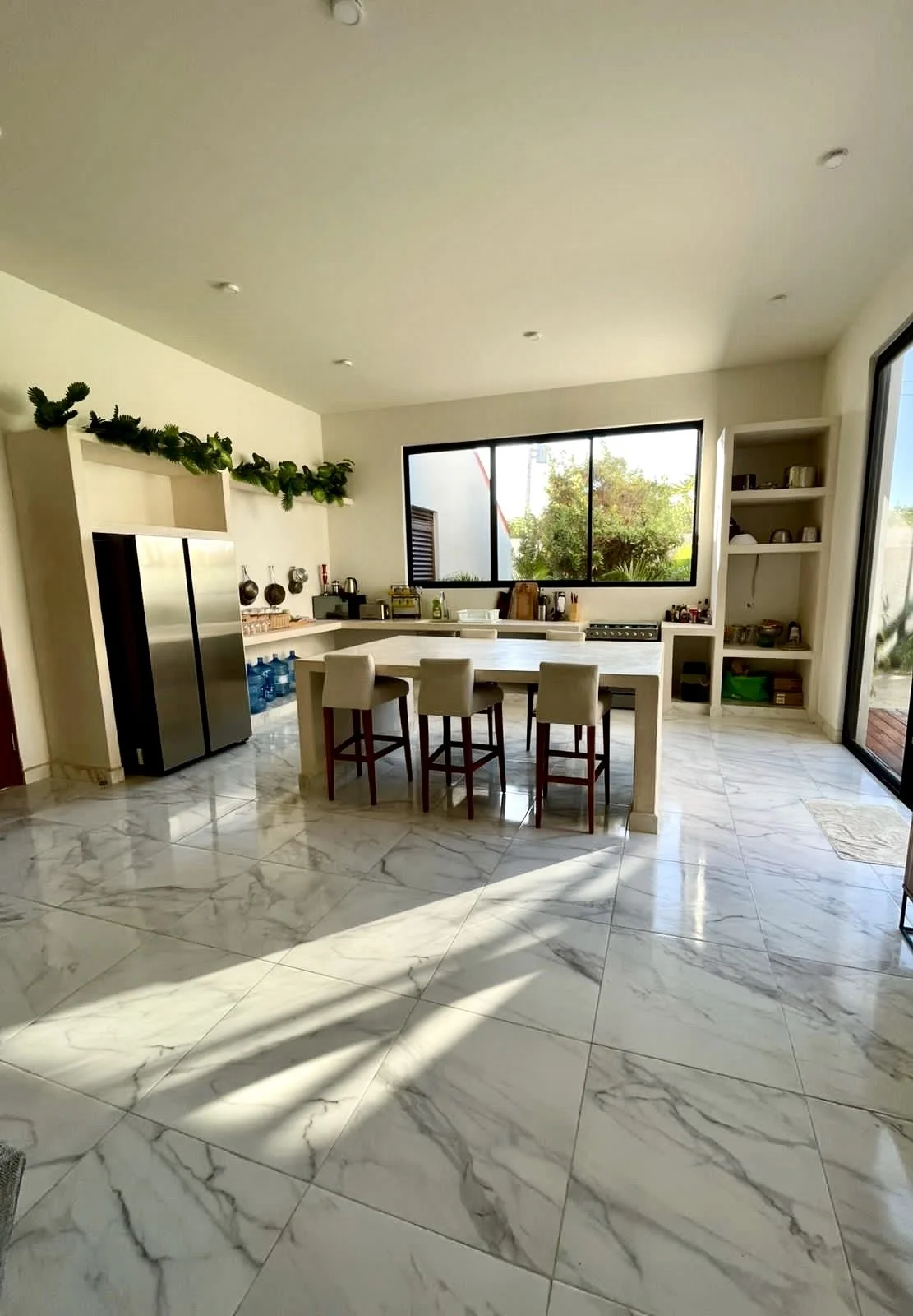 Cuisine lumineuse avec sol en marbre, îlot central avec six chaises, grands fenêtres avec vue sur le jardin, étagères blanches avec vaisselle, réfrigérateur en acier inoxydable.