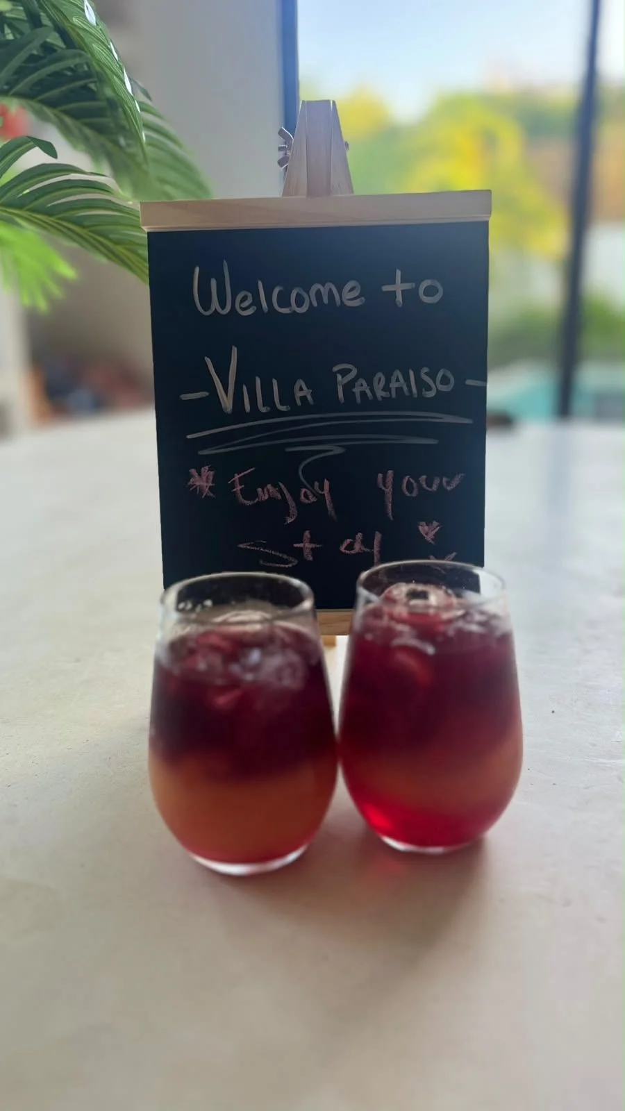 Deux verres de cocktail coloré avec une table à l'avant et un panneau noir derrière indiquant 'Welcome to Villa Paraiso' et une note pour profiter de votre séjour.
