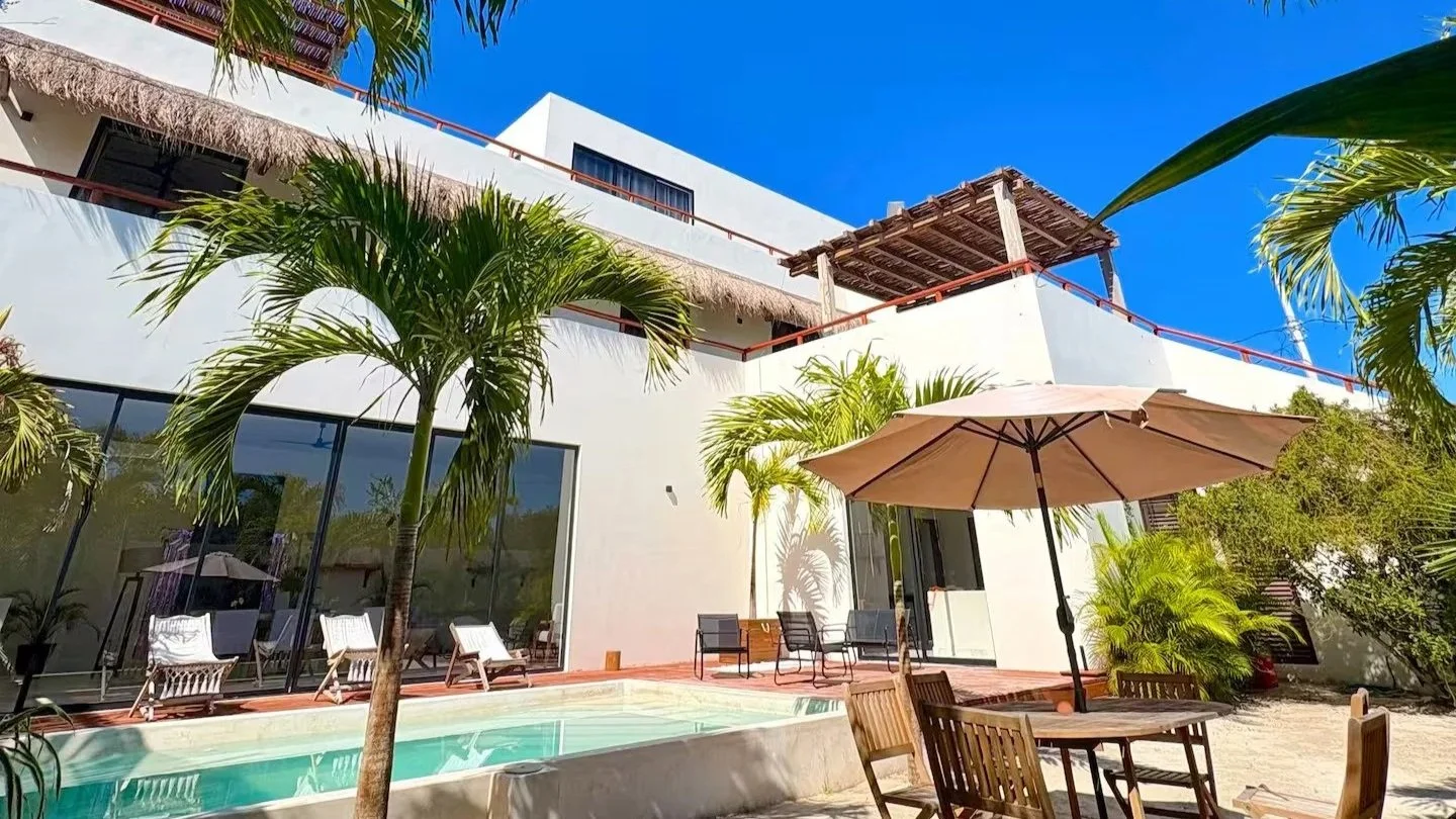 Une villa moderne avec piscine, parasol, chaises en bois, et palmiers en bord de mer, sous un ciel bleu.