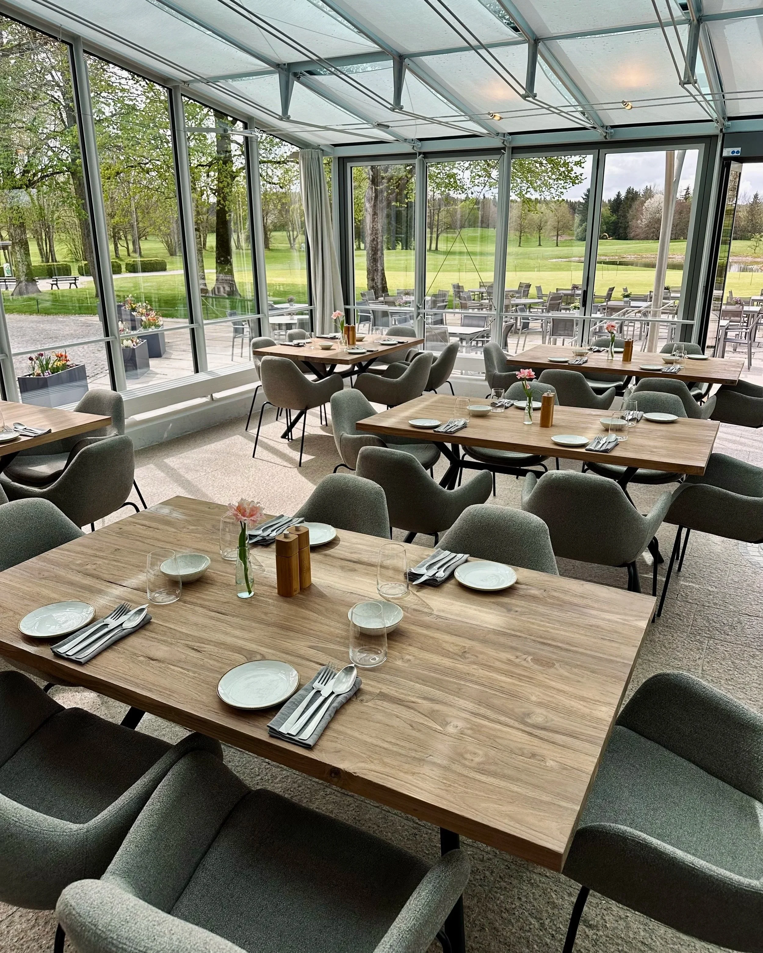 Innenraum eines Restaurants mit Holztischen, Stühlen und einem Blick auf den Garten mit Bäumen im Hintergrund.