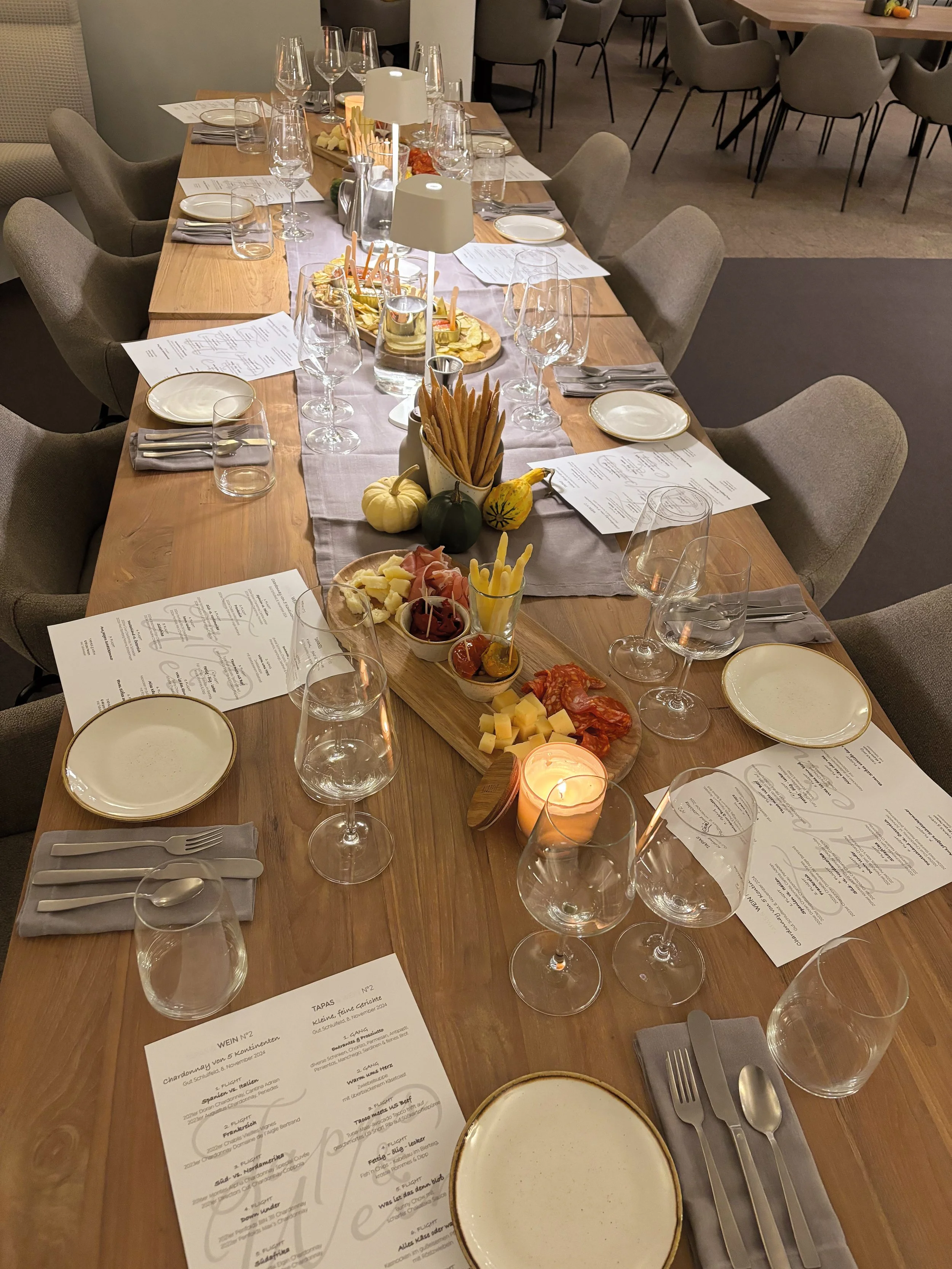 Ein langer Esstisch ist für eine Mahlzeit gedeckt, mit weißen Tellern, Weingläsern, Wasser-gläsern, Messer, Gabeln und Löffeln. In der Mitte des Tisches befinden sich Kerzen, kleine Kürbisse, eine Snackplatte mit Käse, Wurst, und Broten sowie weitere