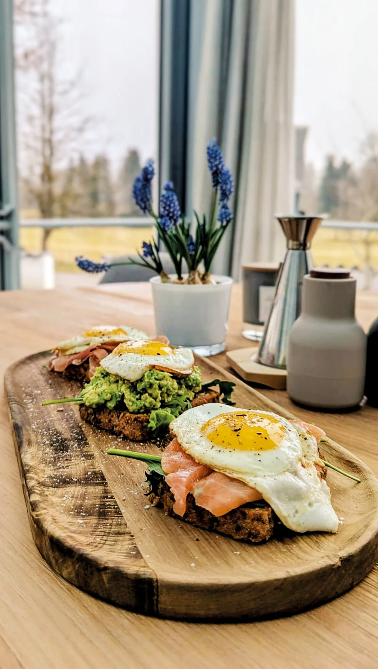 Drei offene Eier und geräucherter Lachs auf Avocado-Toast mit Toastbrot, serviert auf einem Holzbrett.