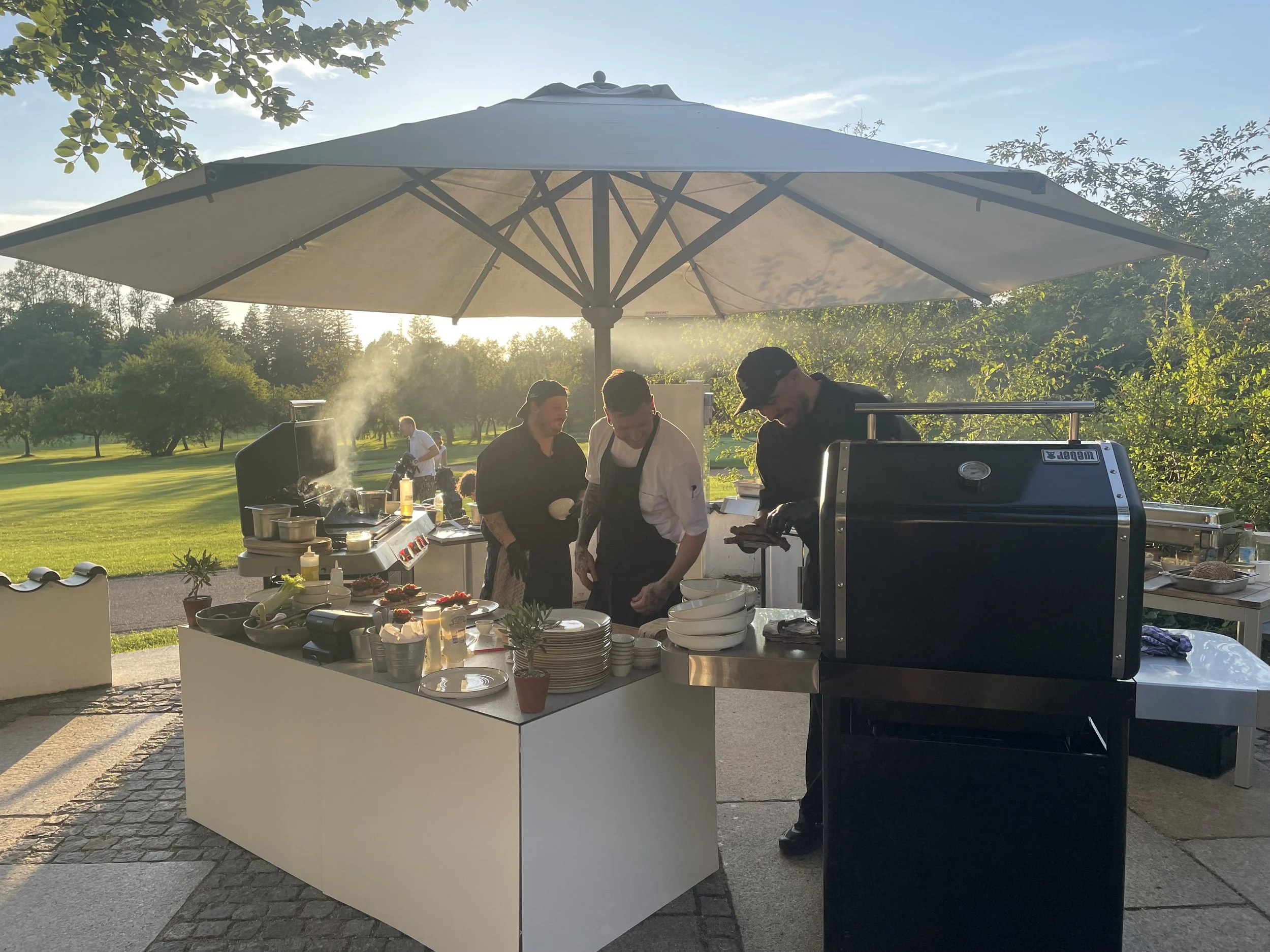 Grillparty im Freien mit mehreren Köchen unter einem großen Sonnenschirm auf einer Wiese, mit verschiedenen Speisen und Kochutensilien auf einem weißen Tisch.