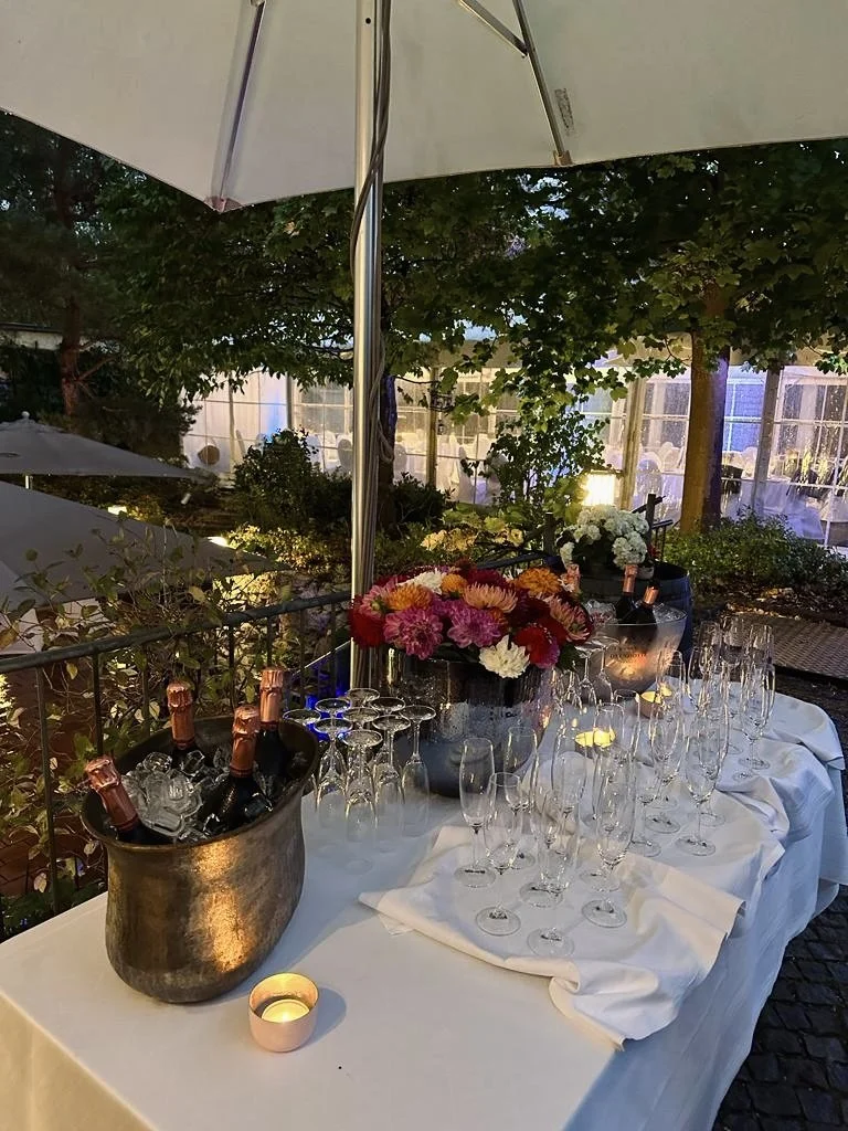 Schöner Getränketisch im Freien mit Blumen, Sektflaschen, Gläser, Kerzen, Sonnenschirm und Hügel im Garten bei Abenddämmerung.