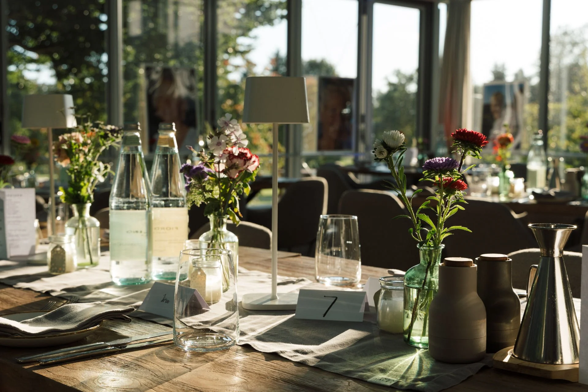 Ein festlich gedeckter Tisch mit bunten Blumen in Vasen, Wasserflaschen, Kerzen, Tischkarten, Geschirr und Besteck in einem hellen Saal mit großen Fenstern, durch die Tageslicht und Außenblick sichtbar sind.