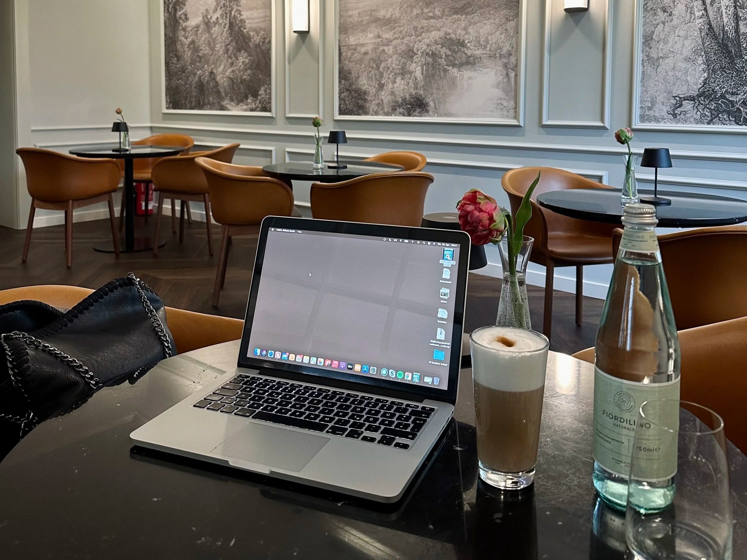 Ein Tisch in einem Café mit einem MacBook, einer Flasche Wasser, einem Latte Macchiato, einer kleinen Blume im Glas und einer schwarzen Tasche.