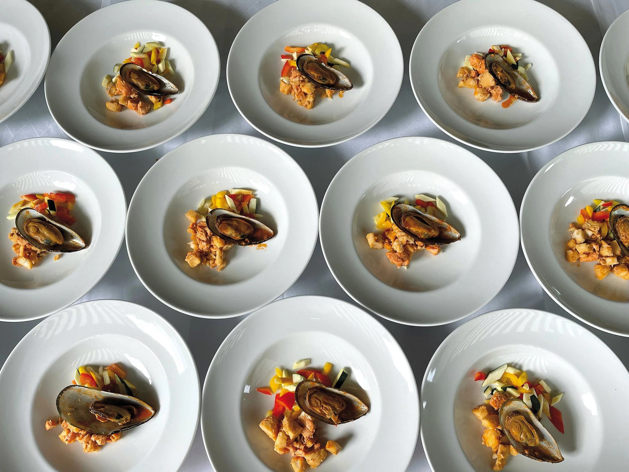 Mehrere weiße Teller mit Muscheln, gemischtem Gemüse und einer Fleisch- oder Fischsauce, ordentlich auf einem Tisch.