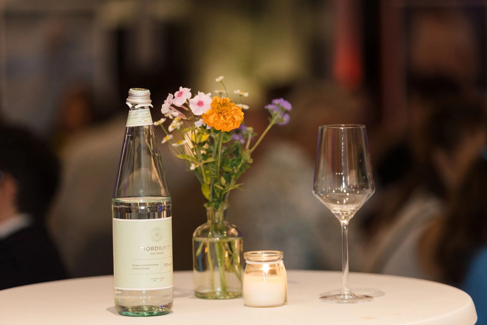 Eine Flasche Mineralwasser, eine Vase mit bunten Blumen, eine Kerze in einem Glas, ein leeres Weinglas auf einem weißen Tisch