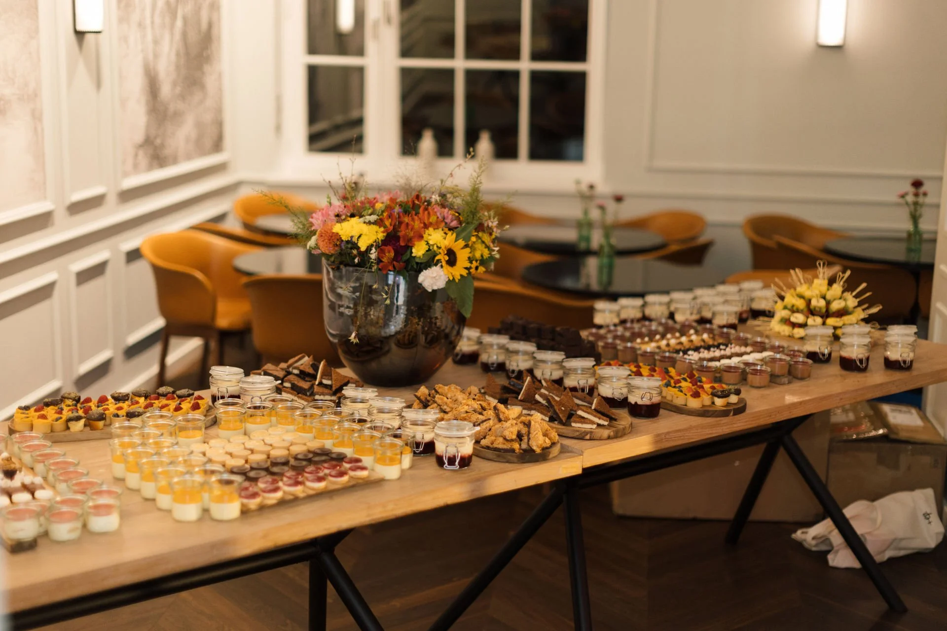 Süßigkeiten und Desserts auf einem Buffet-Table, darunter Gläser mit Pudding, Kuchen, Kekse und Mini-Törtchen, mit einer großen Blumenvasen in der Mitte.