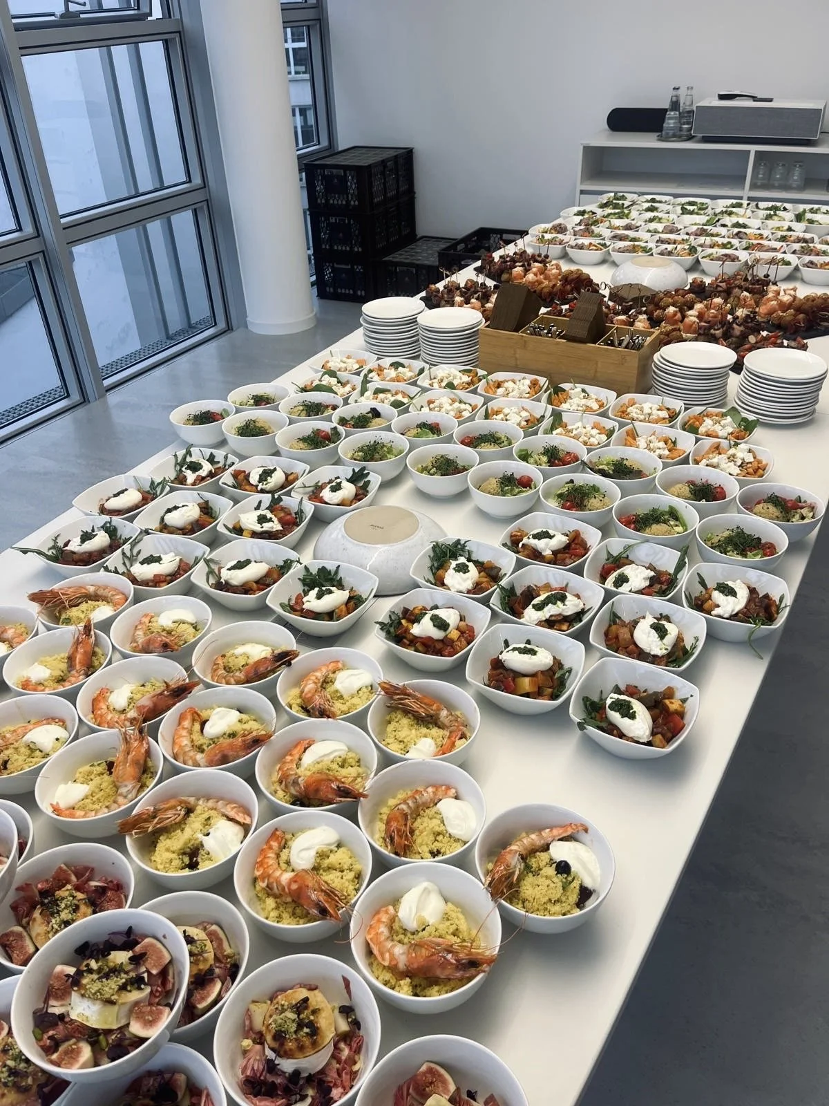 Tisch mit vielen verschiedenen italienischen Gerichten, darunter Garnelengerichte, Salate und Käse, in Weißporzellanschalen in einem hellen, modernen Raum mit großem Fenster.