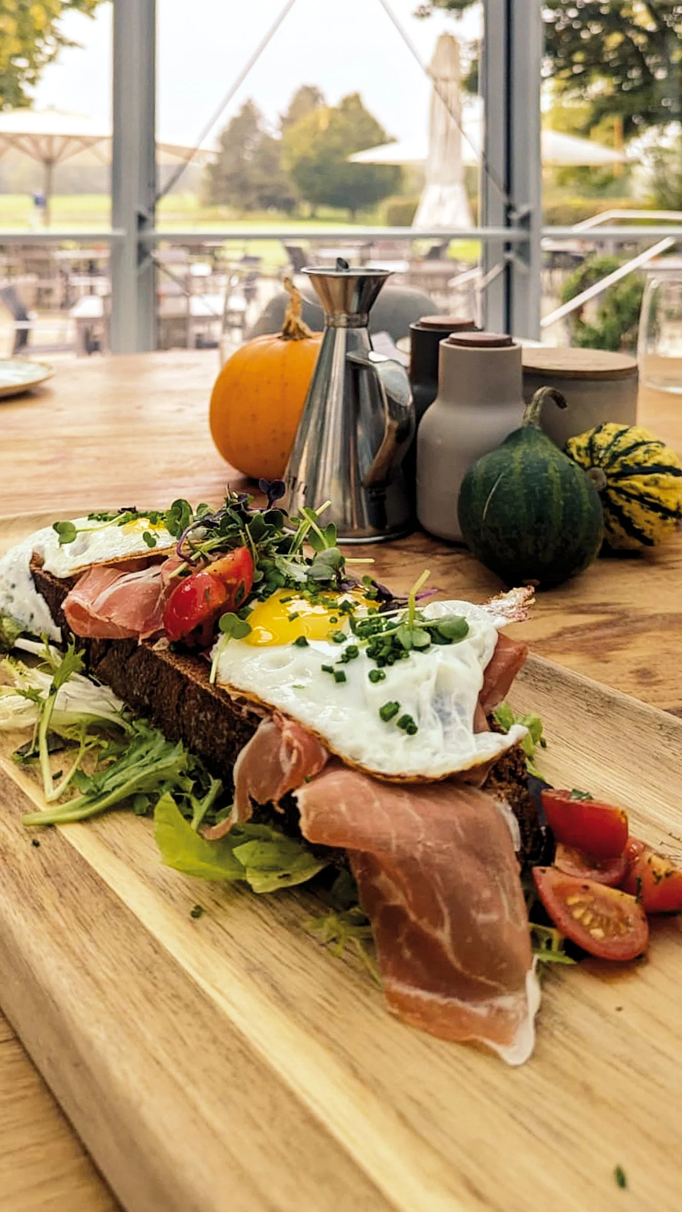 Ein belegtes Brot mit Spiegelei, Schinken, Tomaten und Sprossen auf einem Holzbrett, im Hintergrund Kürbisse, eine Kaffeemaschine und Kaffeekannen auf einem Tisch, mit Blick auf eine grüne Landschaft im Freien.