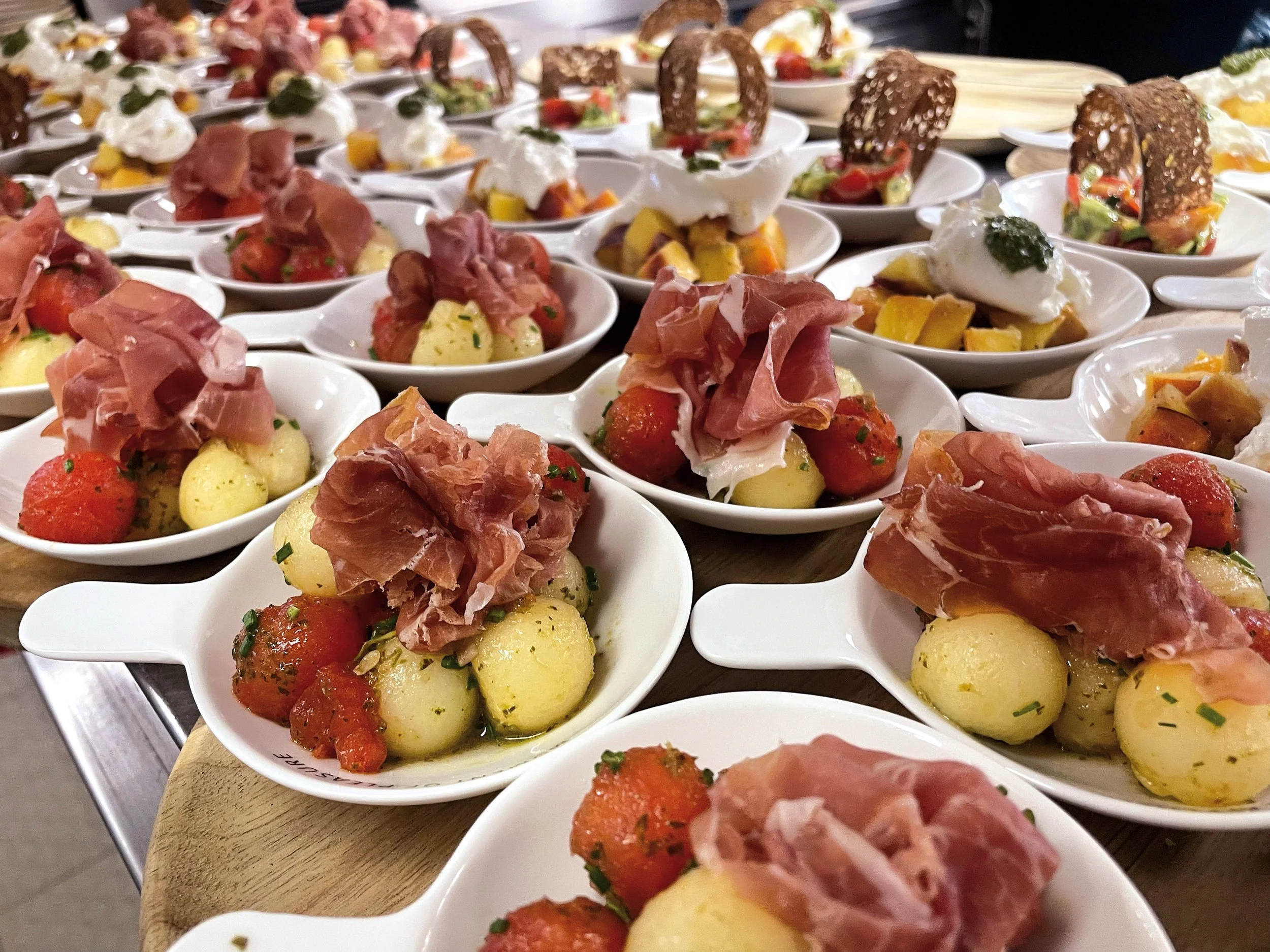 Mehrere kleine weiße Schälchen mit gekochten Kartoffeln, Tomaten, Schinken und Soße auf einem Holztisch.