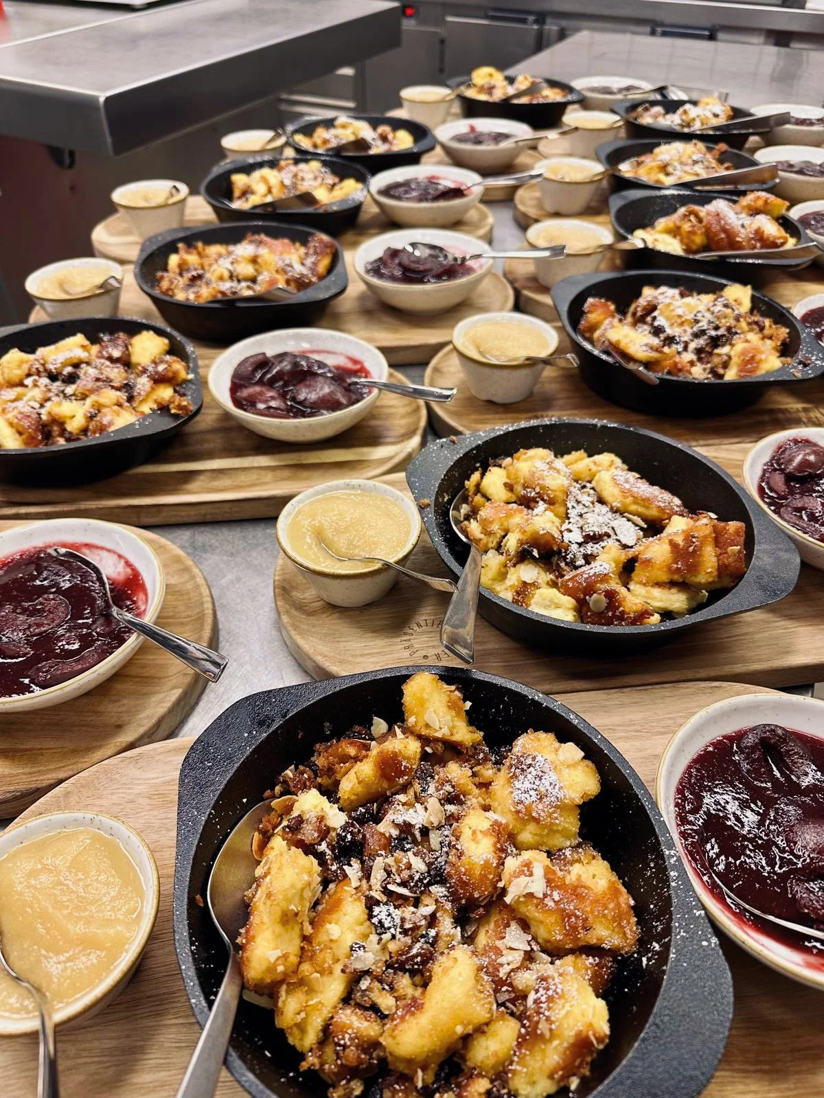 Verschiedene Schalen mit warmer Apfelpfannekuchen, Beeren, und Vanillesoße auf Holzbrettern bei einer Café- oder Frühstücksstation.