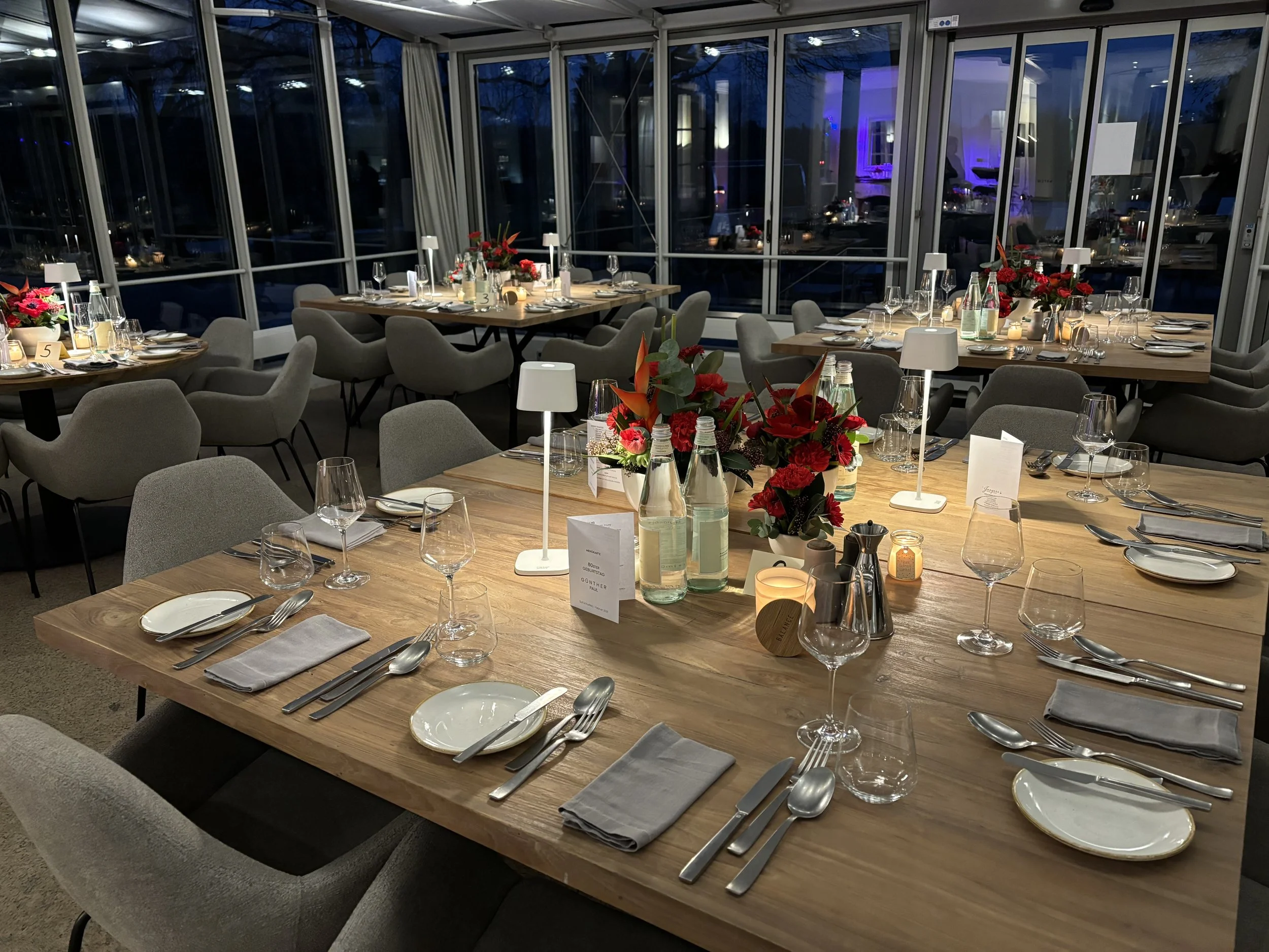 Elegant gedeckter Tisch in einem festlichen Restaurant. Der Tisch ist mit dezenten grauen Stuhlen, weißen Servietten, Silberbesteck, Wein- und Wassergläsern, Kerzen, weißen Lampen, und roten Blumen als Dekorationen ausgestattet. Im Hintergrund sind g