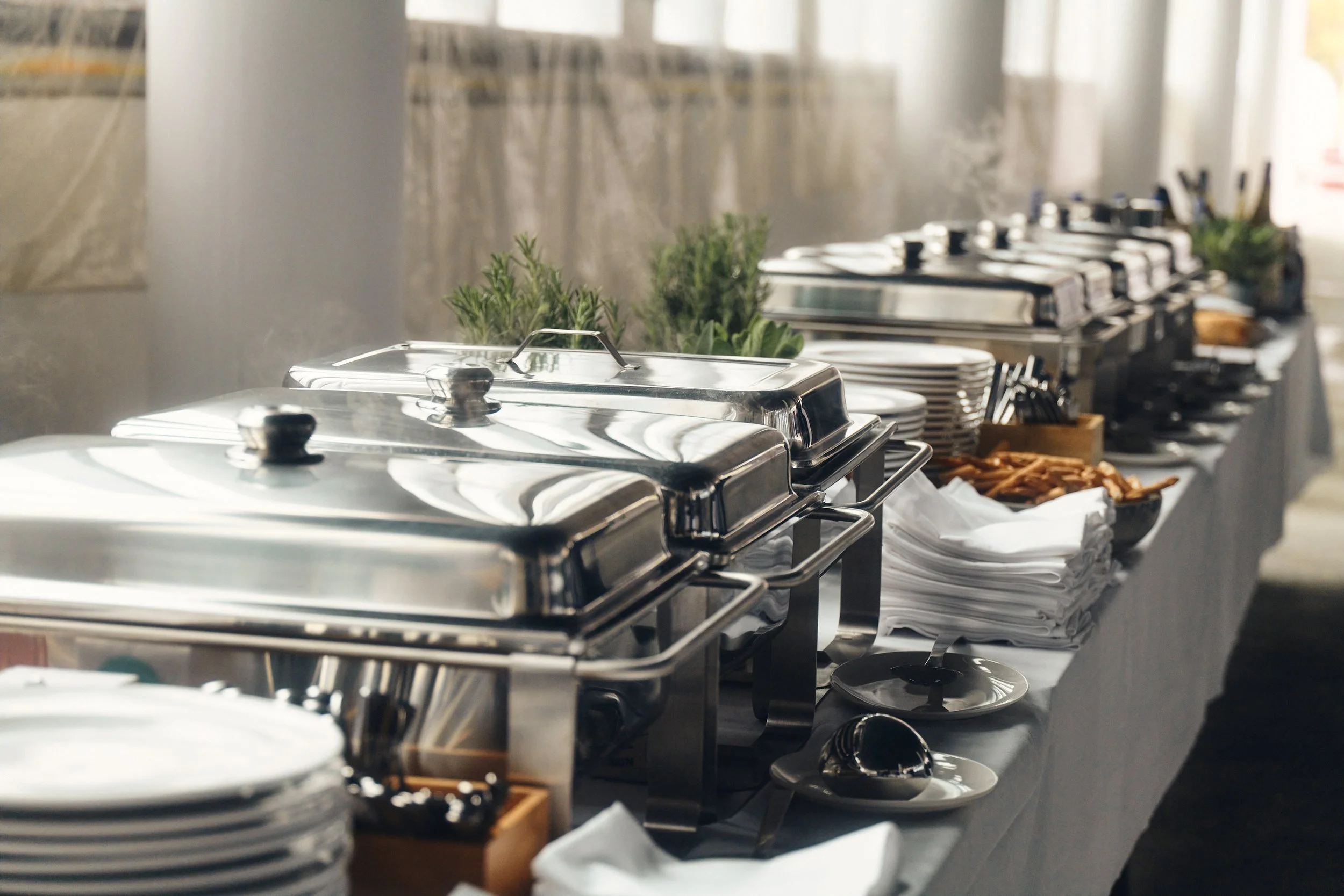 Buffet-Tisch mit mehreren Edelstahl-Wärmebehältern, Tellern, Besteck, frischen Kräutern und Snacks bei einer Veranstaltung.