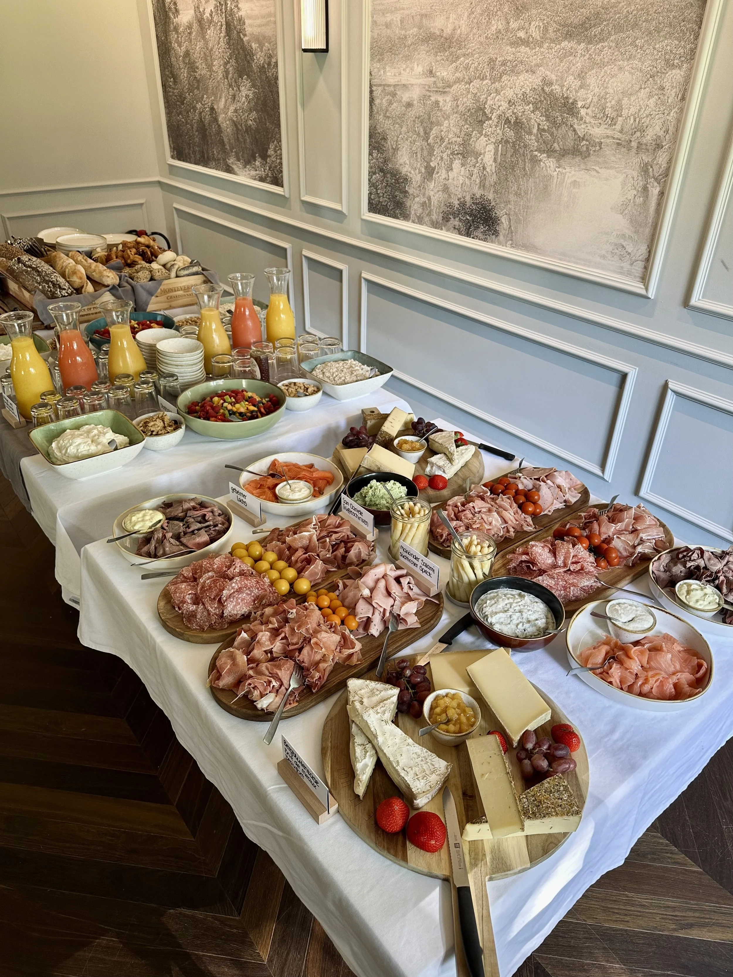 Ein Frühstücksbuffet mit Käse, Aufschnitt, Früchten, Joghurt, Säften und Backwaren auf einem langen Tisch mit weißem Tischtuch.