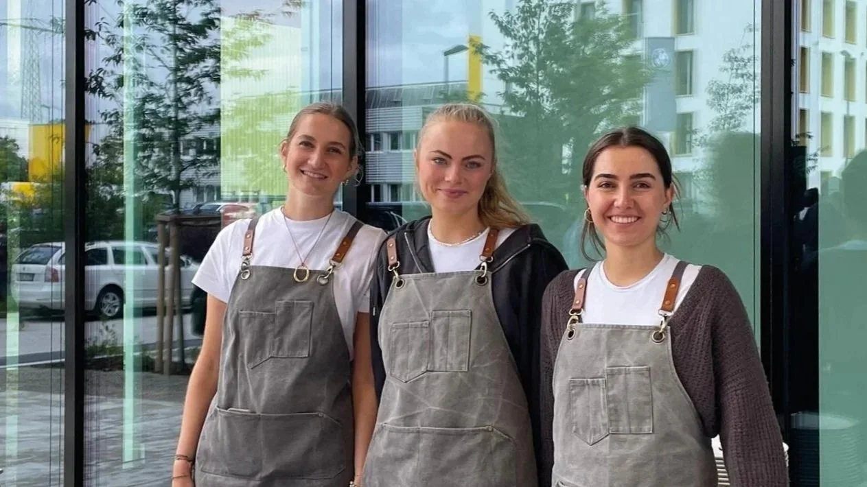 Drei junge Frauen stehen vor einem Glasfenster, tragen graue Schürzen und lächeln in die Kamera.