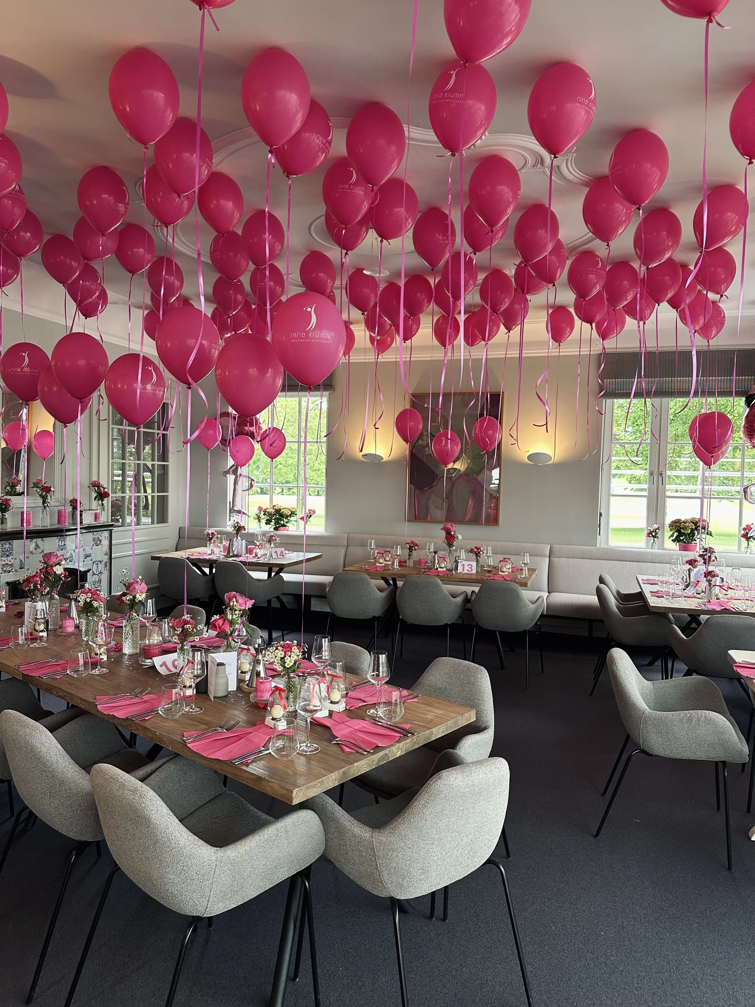 Ein festlich dekorierter Raum mit vielen pinken Ballons an der Decke, Tischen mit pinken Servietten, Blumen und Gläsern.