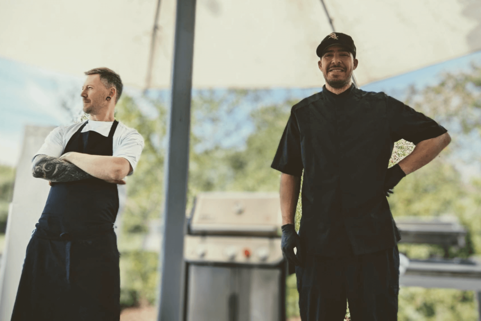 Zwei männliche Köche im Freien, einer trägt ein weißes T-Shirt und eine schwarze Schürze, der andere trägt eine schwarze Uniform und eine schwarze Mütze, lächelnd, mit einem Grill im Hintergrund.