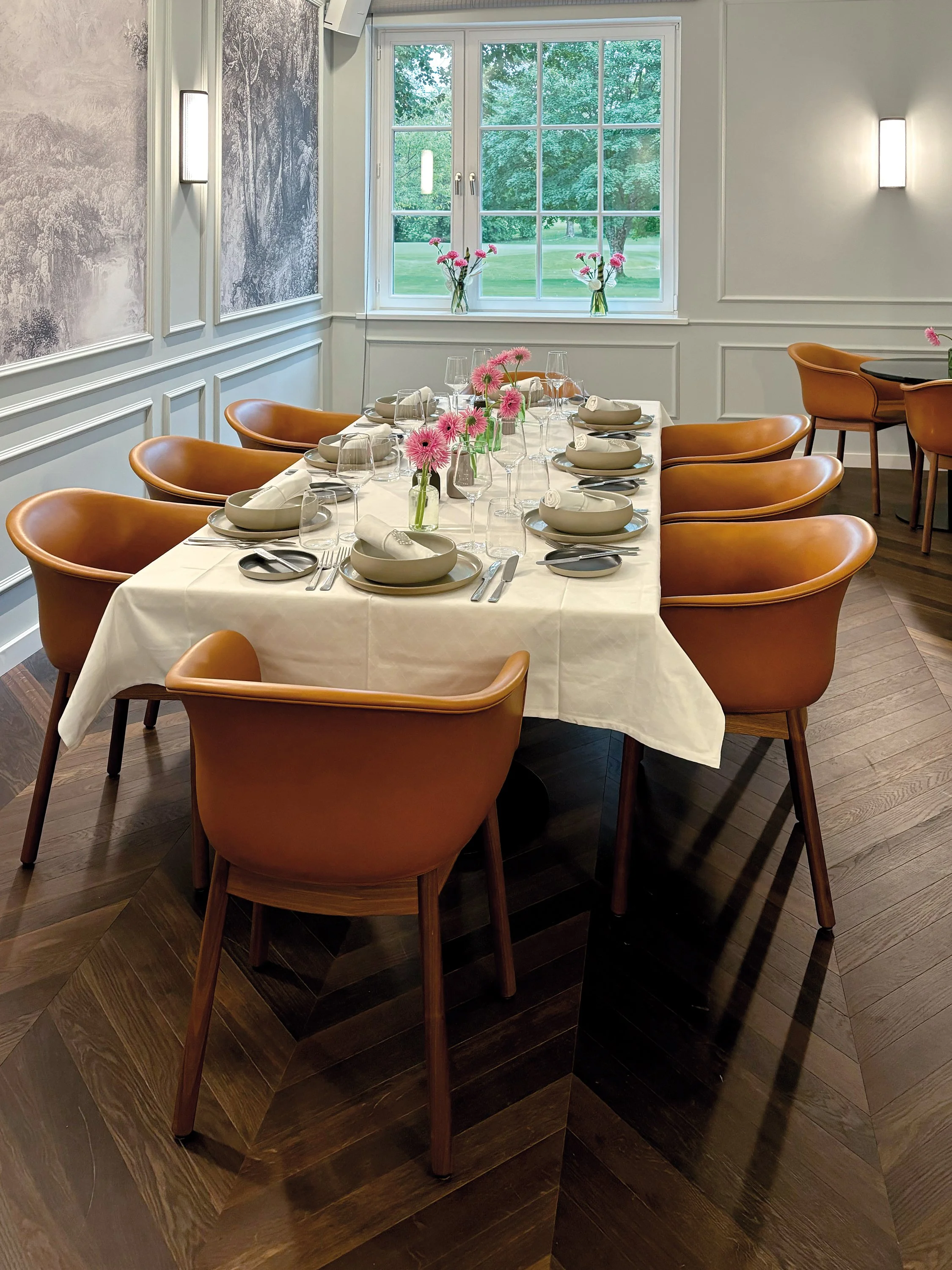 Ein festlich gedeckter Esstisch mit weißen Tischtuch, weißen Tellern, Gläsern und Besteck, dekoriert mit pinken Blumen in Gläsern, in einem hell erleuchteten Raum mit großen Fenstern, einem Blick auf einen grünen Garten.