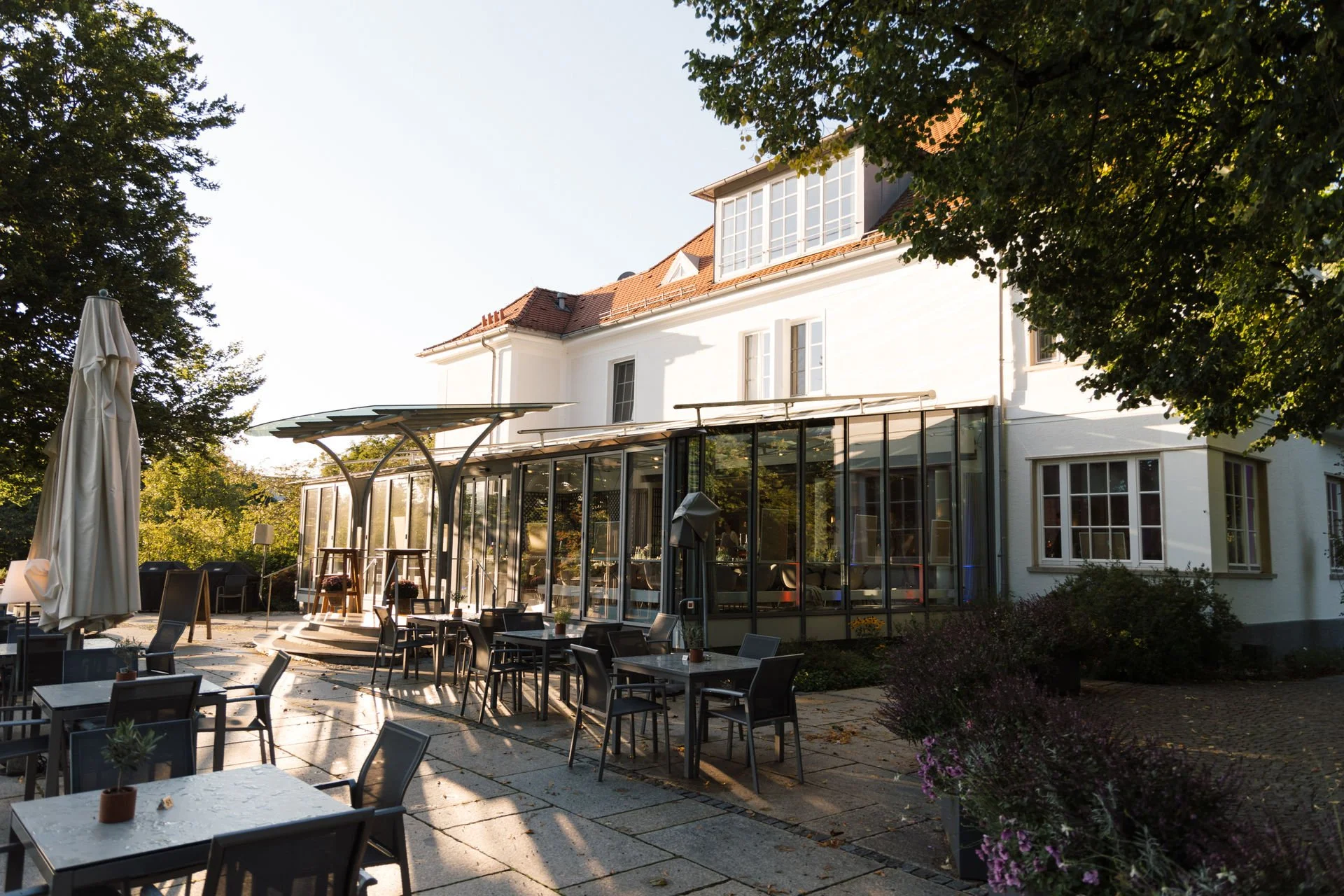 Außerhalb eines Restaurants oder Cafés im Garten sitzen Tische und Stühle, ein Sonnenschirm und eine Terrasse, viele Bäume und ein weißes Gebäude im Hintergrund bei Sonnenuntergang.