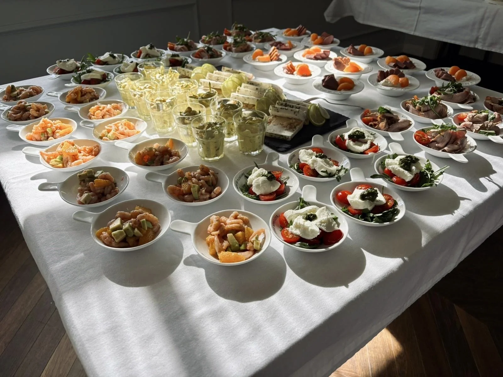 Verschiedene Vorspeisen und Häppchen auf einem Esstisch, darunter Caprese-Salat, Tomaten mit Mozzarella, Antipasti, kalte Platten und kleine Gläser mit Getränken, auf weißem Tisch. Sonnenlicht fällt auf die Speisen.