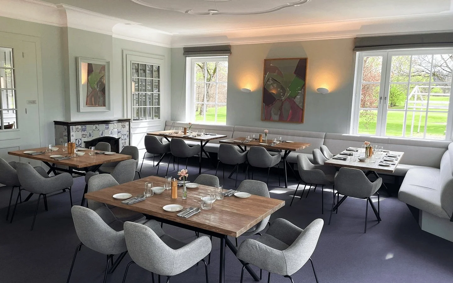 Eleganter Essraum mit mehreren Holztischen, grauen Stühlen, großen Fenstern, Kunstwerken an der Wand, einem Kamin, und Blick ins Grüne draußen.