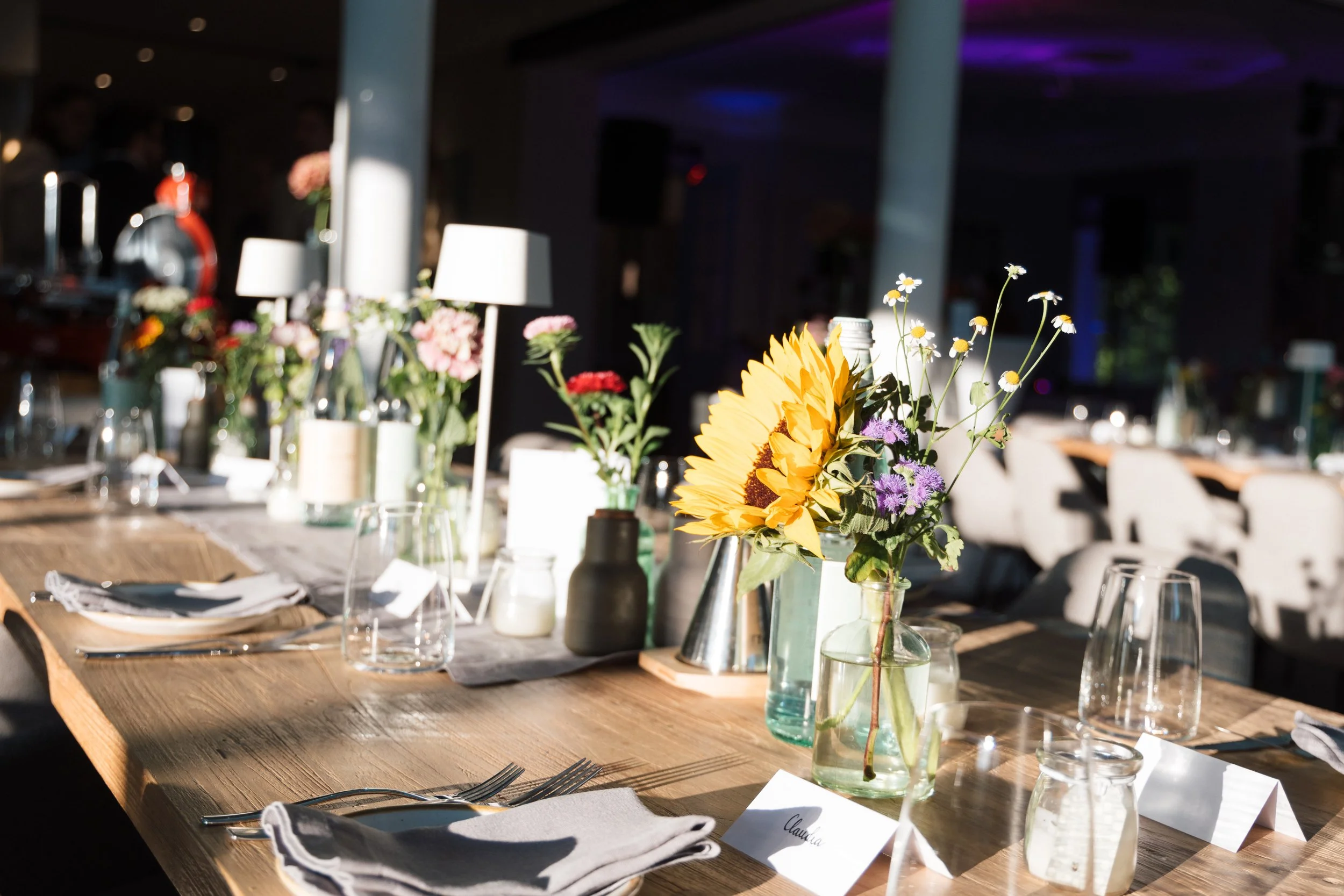 Ein gedeckter Tisch mit Blumen in Gläsern, Tellern, Besteck, und kleinen Lampen, bei Tageslicht in einem festlich dekorierten Raum.