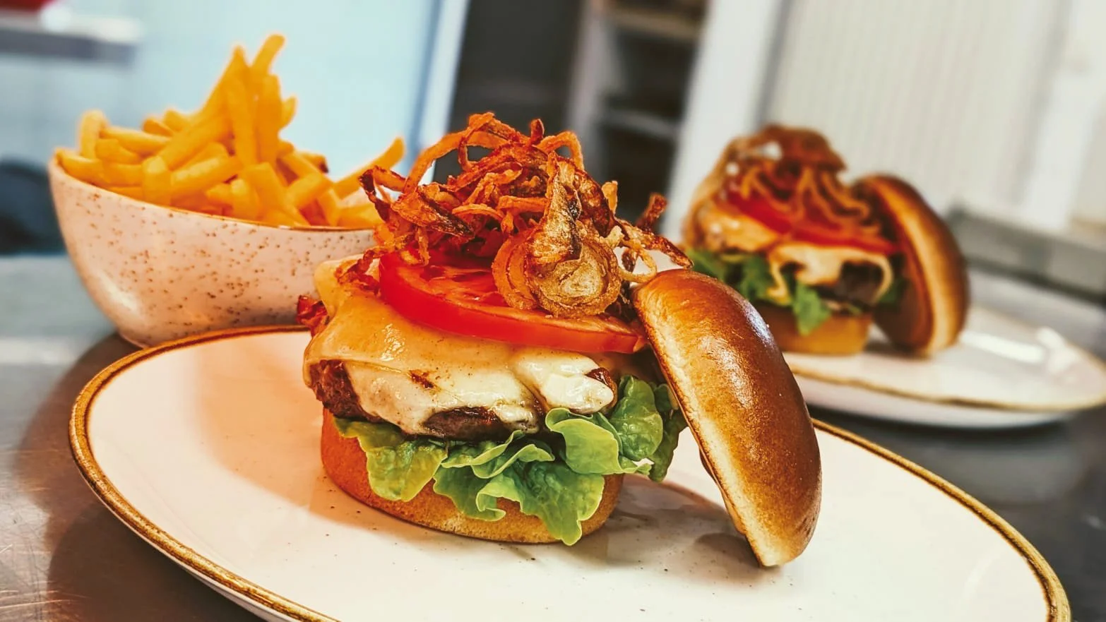 Ein Burger mit Fleisch, Käse, Tomate, Salat und Zwiebelrösti auf einem weißen Teller, dazu eine Schüssel Pommes frites.