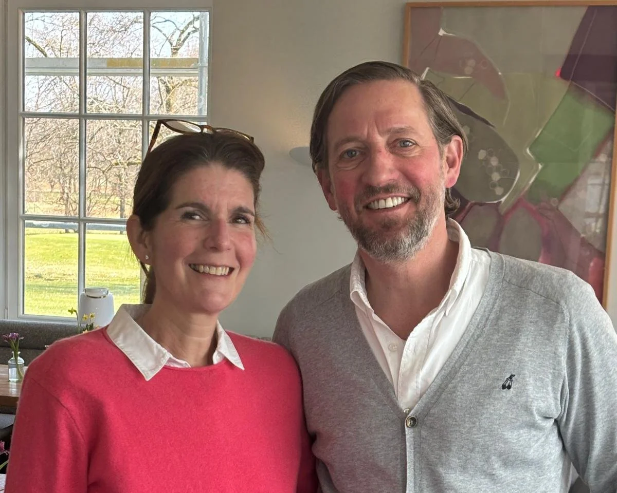 Ein lächelndes Paar steht nebeneinander in einem hellen Raum. Im Hintergrund sind große Fenster mit Blick auf einen grünen Garten und Bäume, sowie ein modernes Gemälde an der Wand.