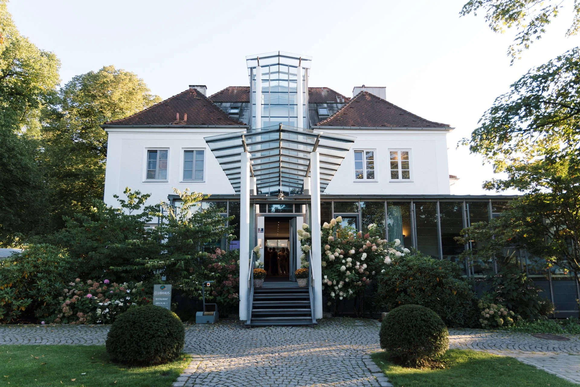 Ein mehrstöckiges, weißes Gebäude mit schrägem Dach, umgeben von Büschen und Blumen. Es gibt eine Treppe mit weißen Stützpfeilern und eine moderne Glasüberdachung am Eingang.