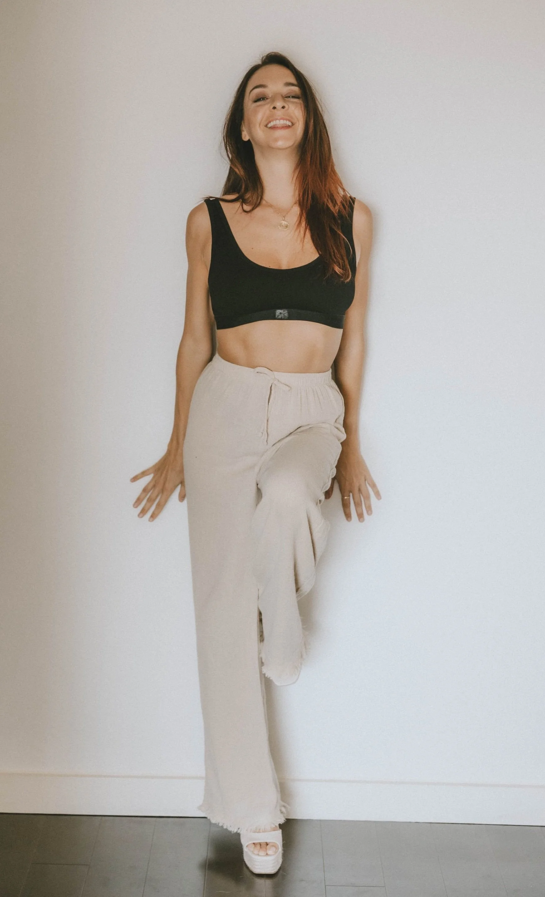 A young woman with brown hair and light skin, smiling while standing against a plain white wall, wearing a black crop top and beige wide-leg pants.