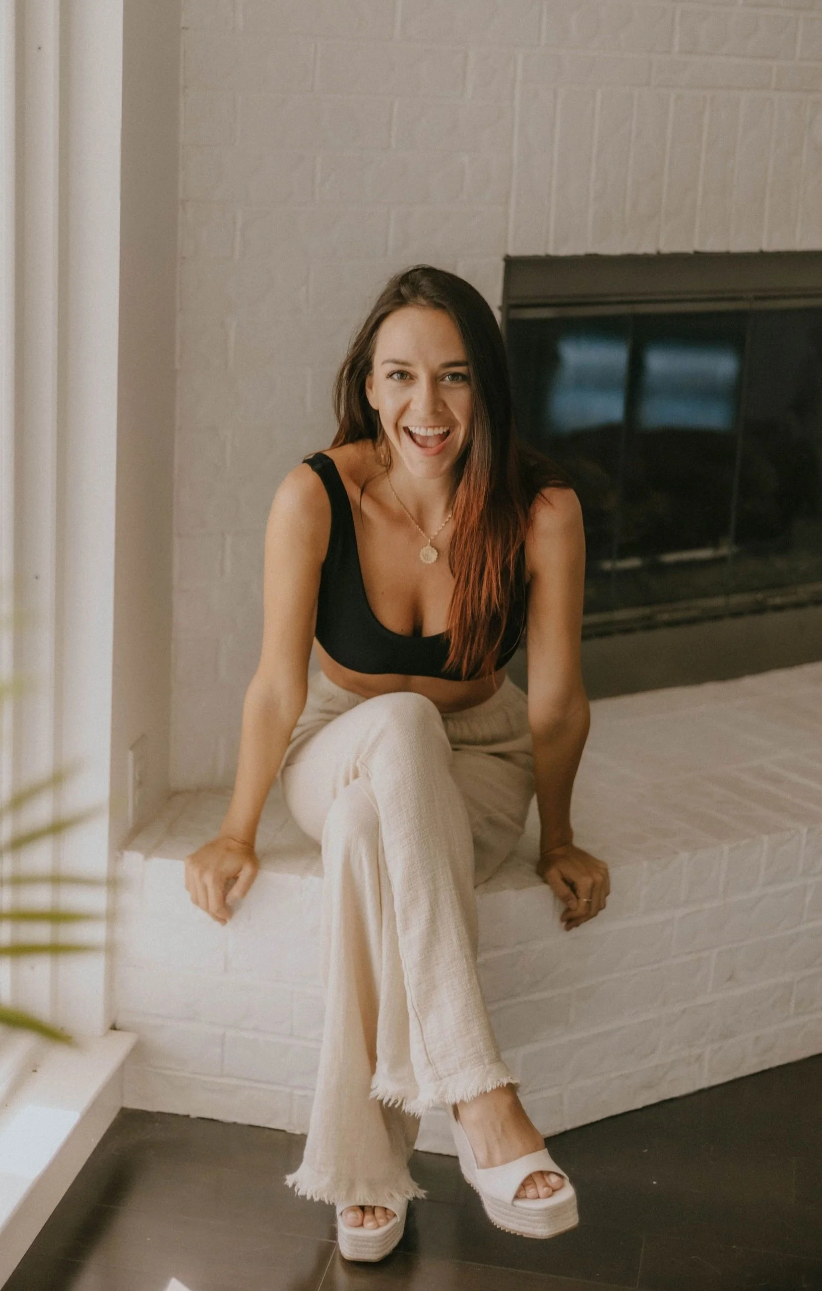 A young woman with long brown hair and a black crop top sitting on a white brick fireplace, smiling and sticking out her tongue, wearing beige wide-leg pants and white platform heels.