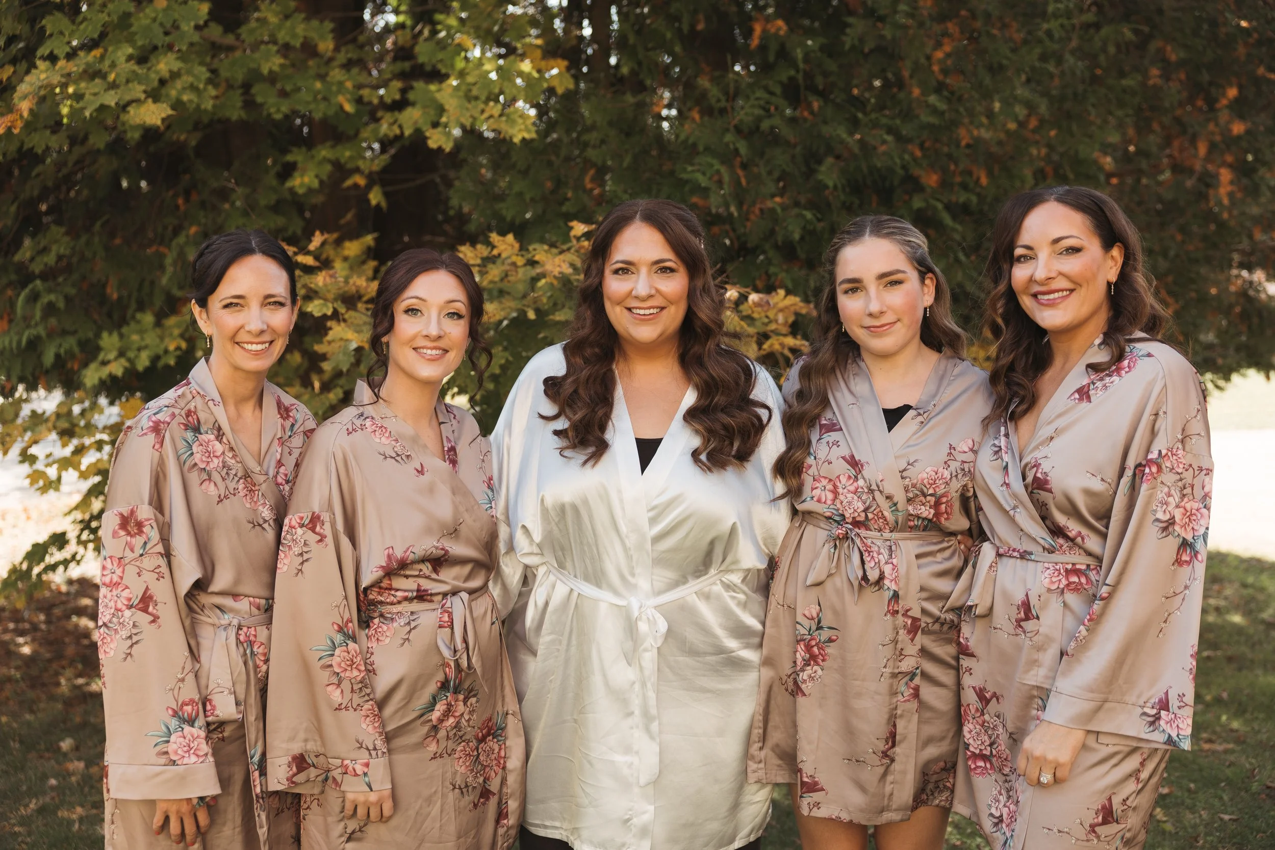 Group of six women outdoors, wearing matching floral robes, posing in front of trees with autumn leaves.