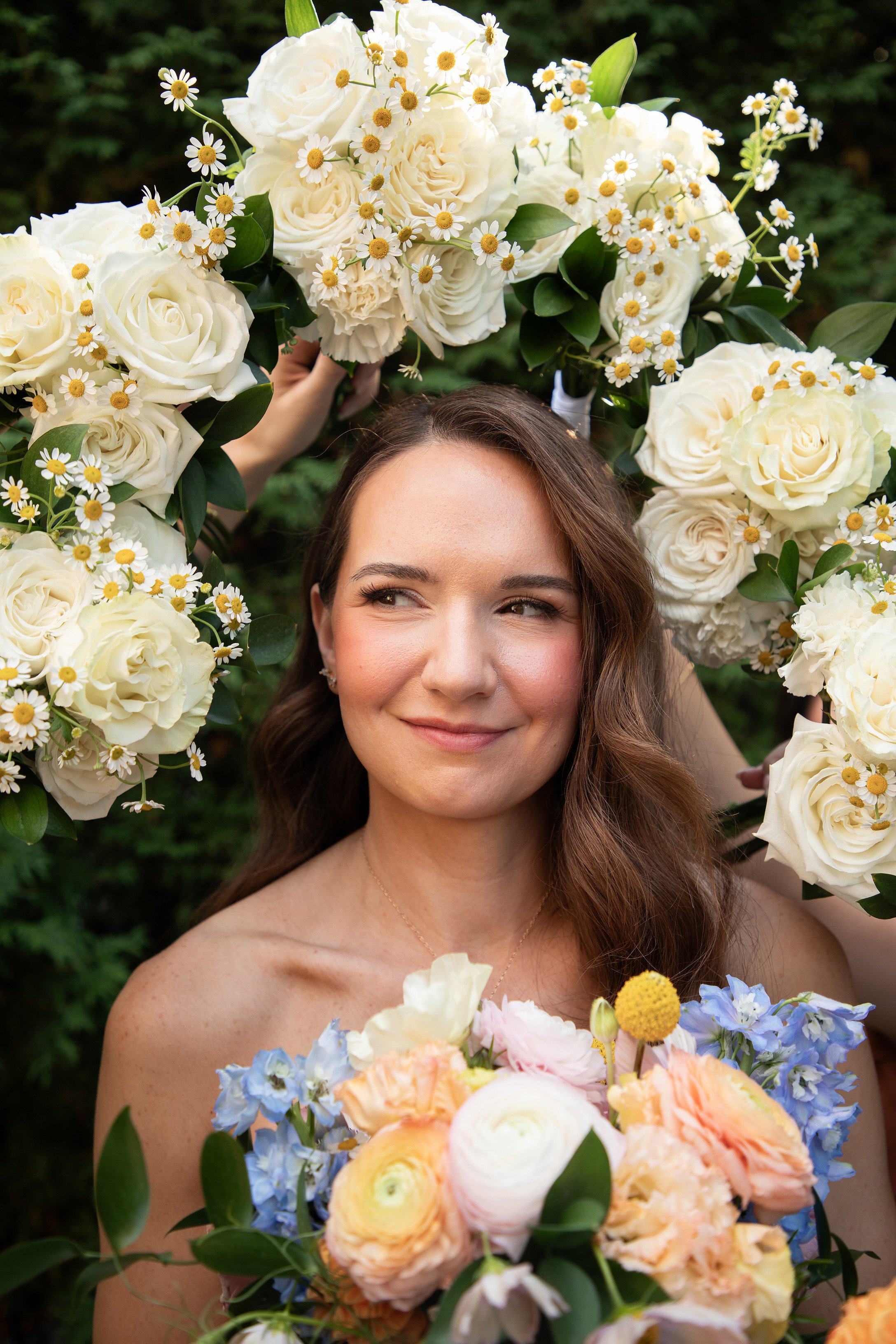 A woman with long brown hair and light skin, smiling softly, surrounded by and holding colorful floral bouquets and a floral wreath around her head, with a dark green leafy background.