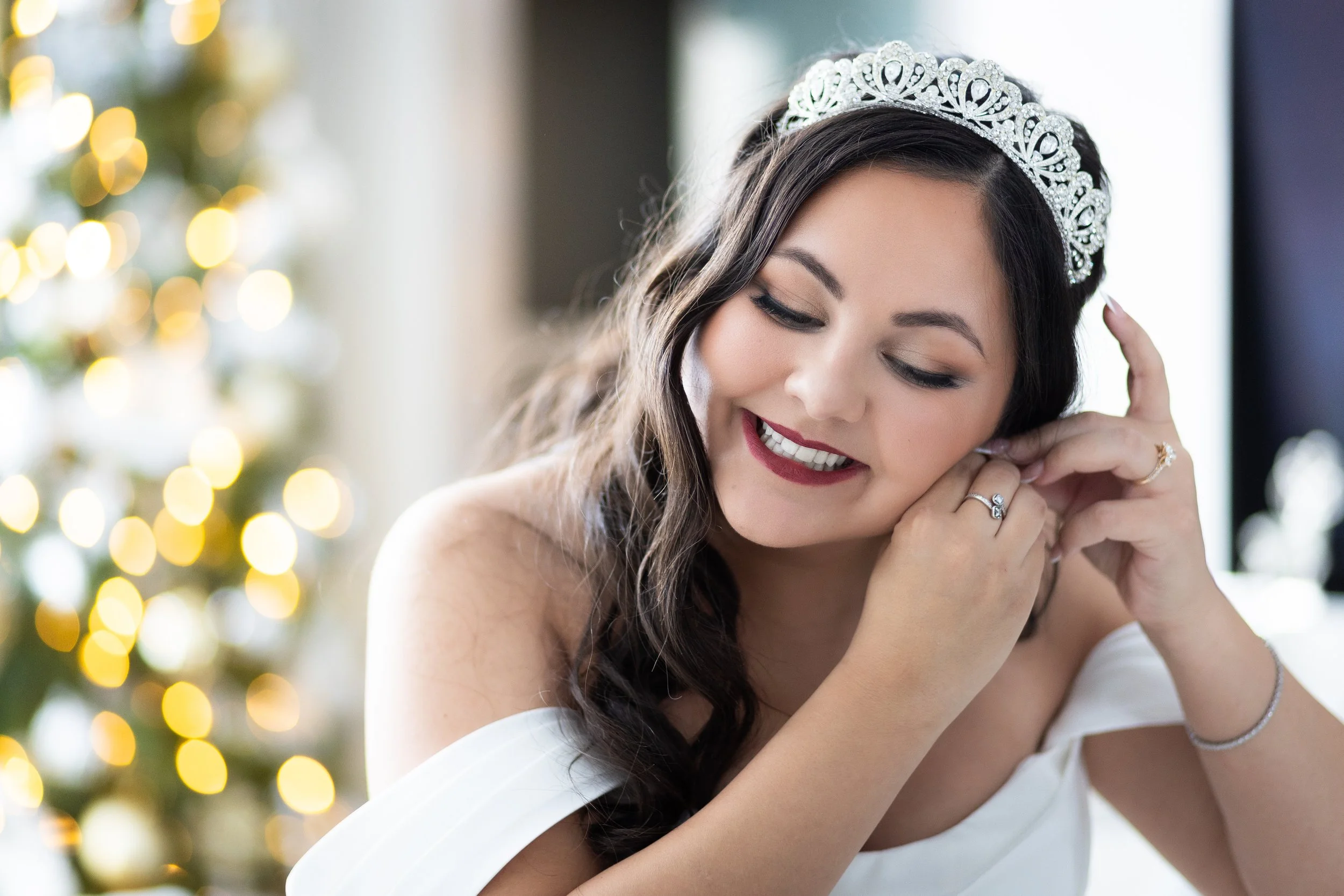 A woman with long dark hair wearing a white gown and a tiara, smiling and adjusting her earring, with a blurred Christmas tree with lights in the background.