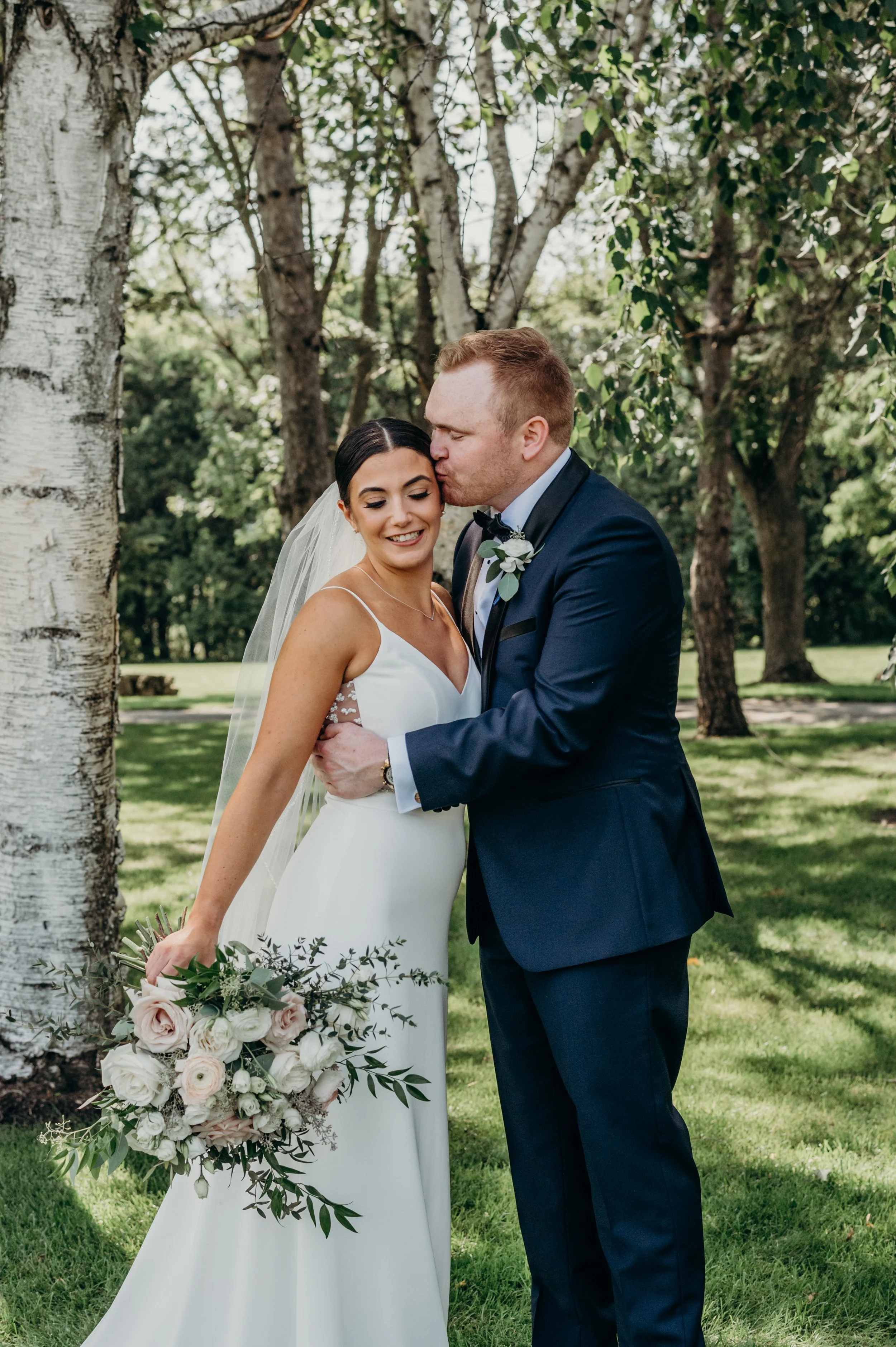 A bride and groom standing close together outdoors, with the groom tenderly kissing the bride's forehead, surrounded by green trees.